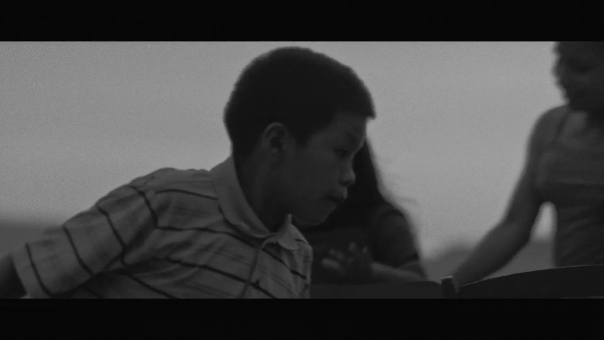 A young boy in a striped polo shirt looks down, while a blurry girl is visible in the background, in a black and white shot.