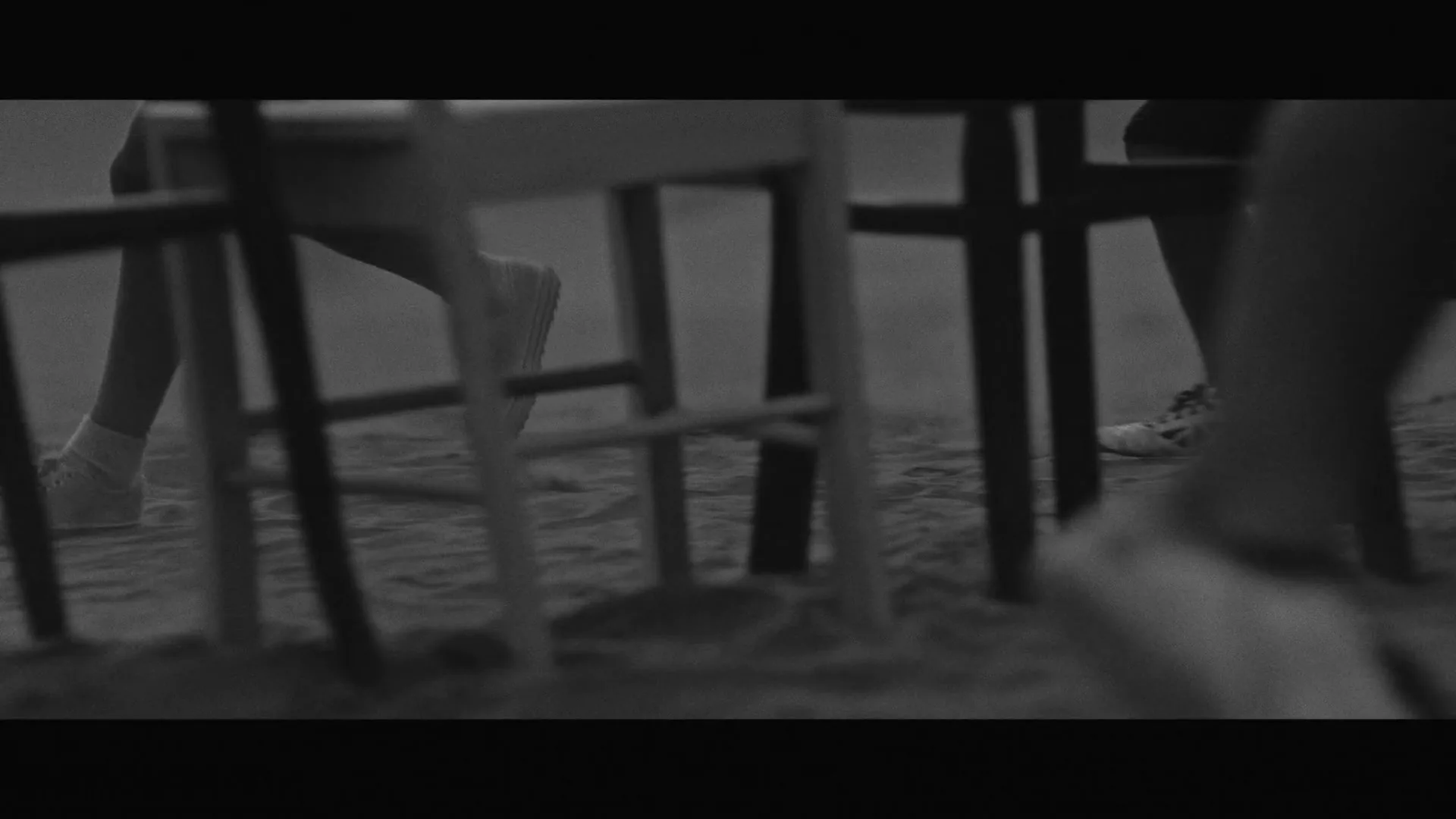 Feet of people in sneakers and bare feet are seen moving around chairs stuck in the sand, in a low-angle, black and white shot.
