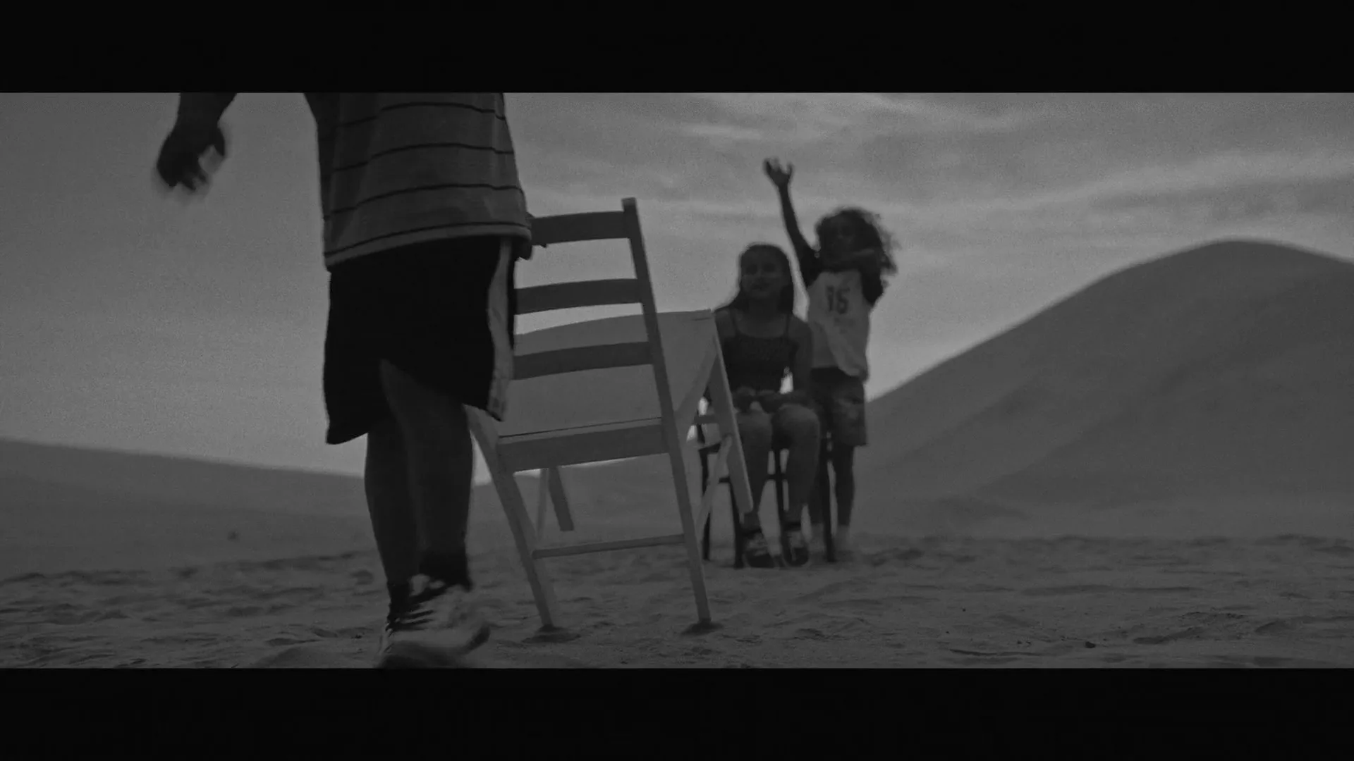 A group of children play musical chairs in a vast, sandy desert landscape under a cloudy sky, with one girl smiling at the camera.
