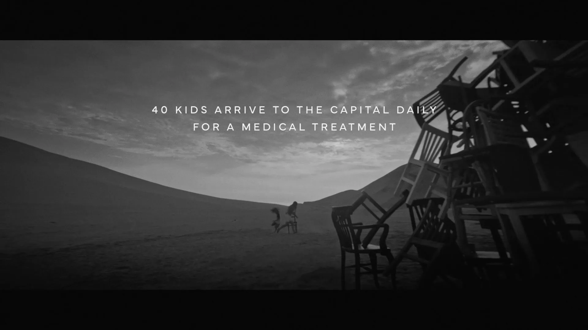 Two children, a girl and a boy, are seen in a desolate, mountainous landscape, pushing a single chair, then more chairs appear, forming a large pile.