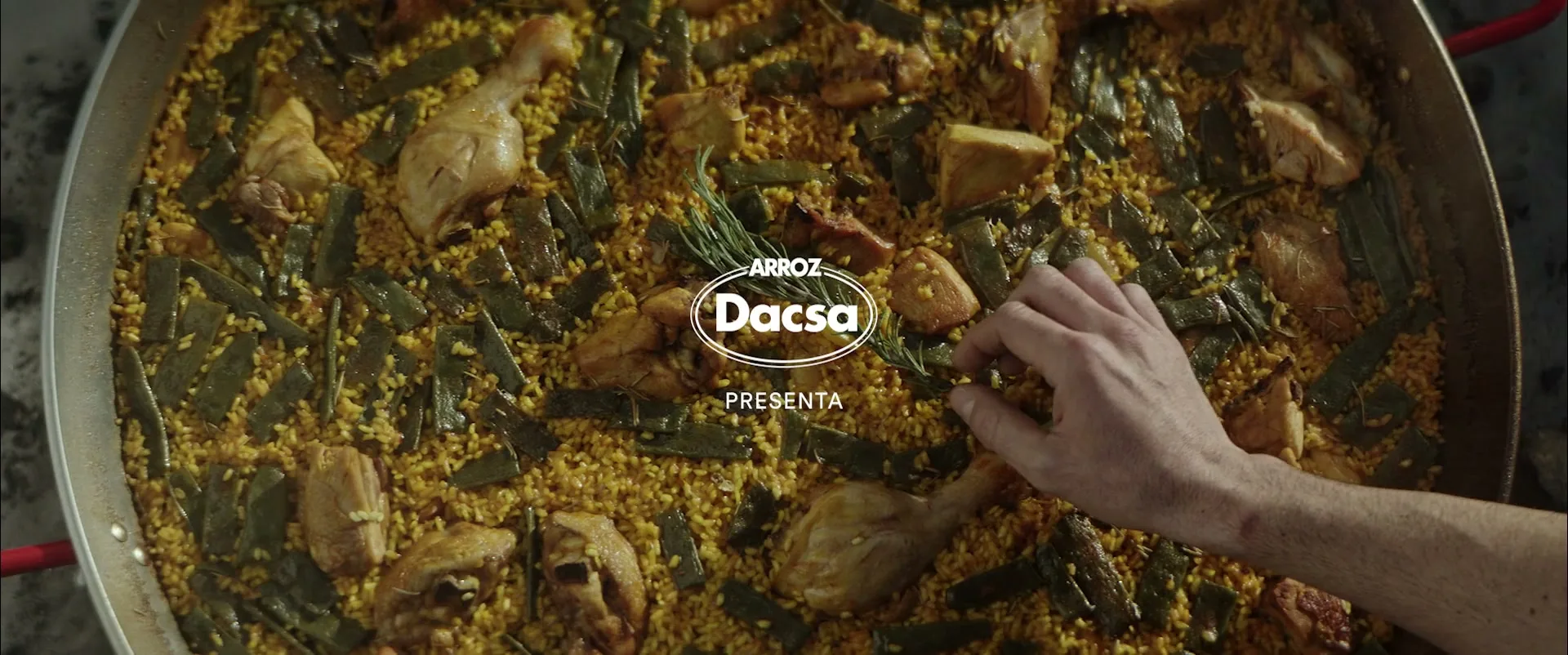 A close-up overhead shot shows a hand placing a sprig of rosemary into a large pan of steaming paella with chicken and green beans.