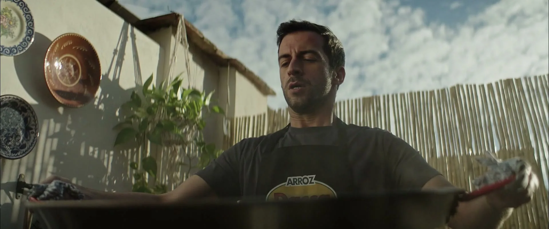 A man in his 30s, wearing a black apron, cooks paella in a large pan on an outdoor patio under a bright sky.