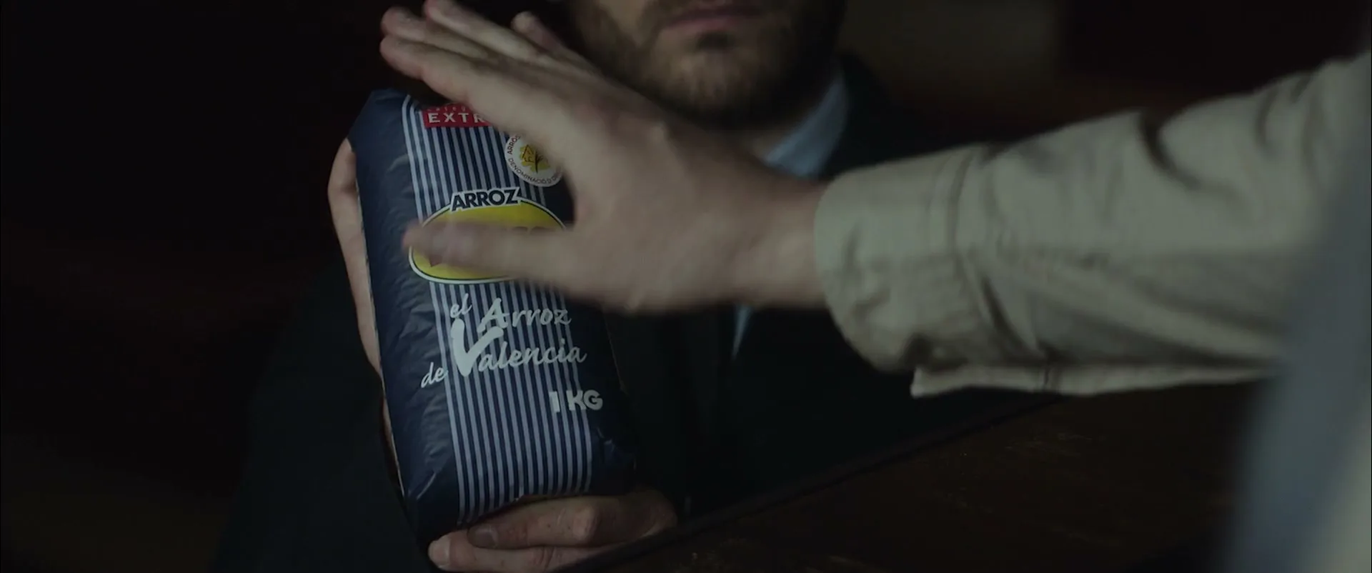 A man in a suit holds a bag of Dacsa rice as another person's hand reaches out to touch it, in a dimly lit interior.
