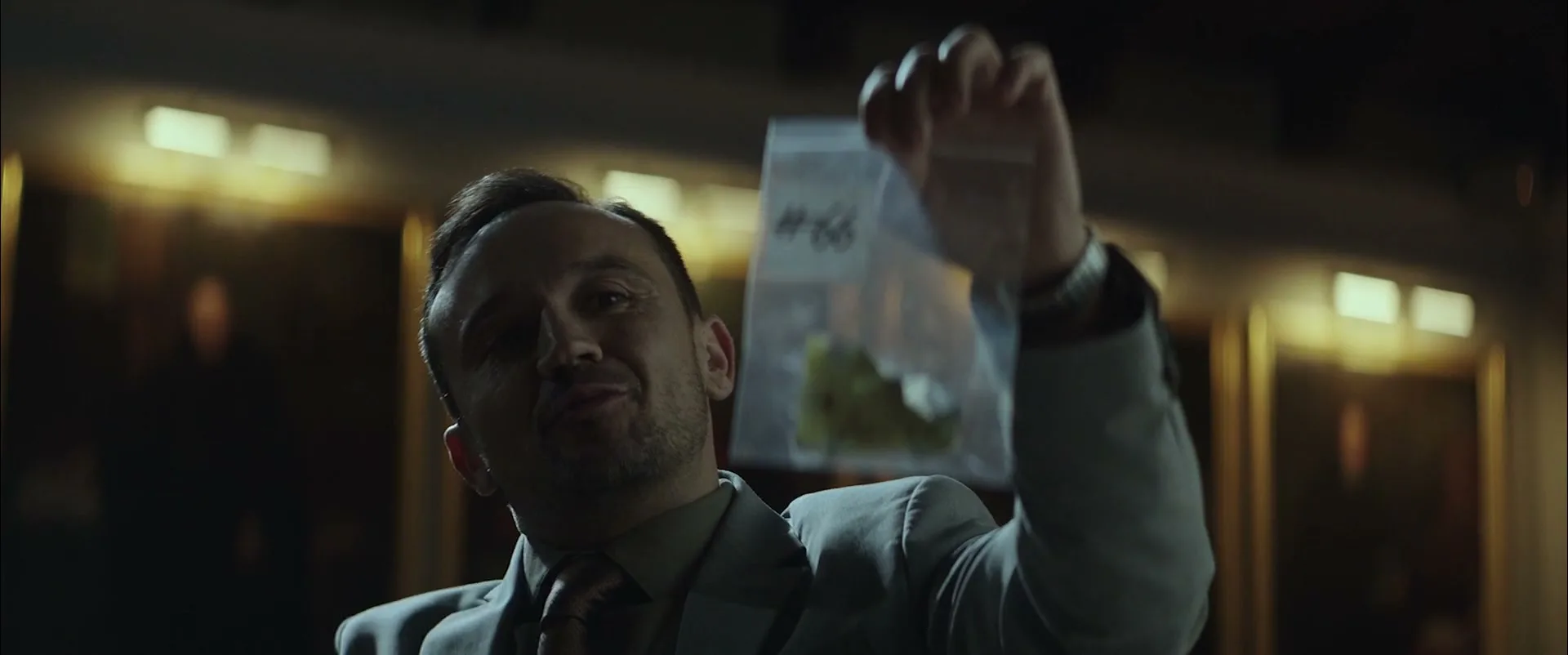 A man in a suit holds up a small plastic bag with green and white contents, looking intently at it in a dimly lit, ornate room.