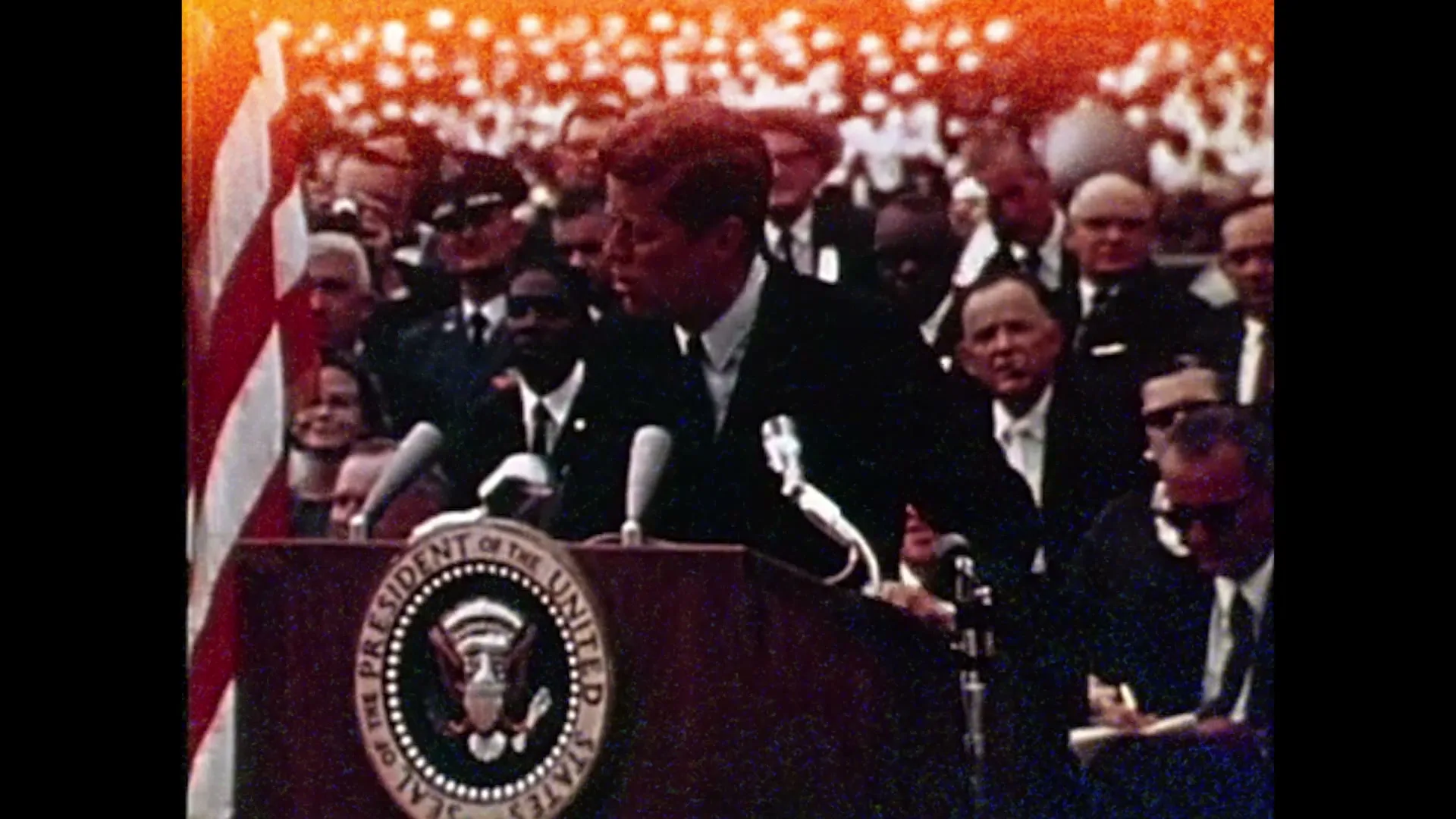 John F. Kennedy, a male in his 40s, wearing a suit, speaks at a podium with the Presidential Seal, addressing a crowd outdoors during the day.