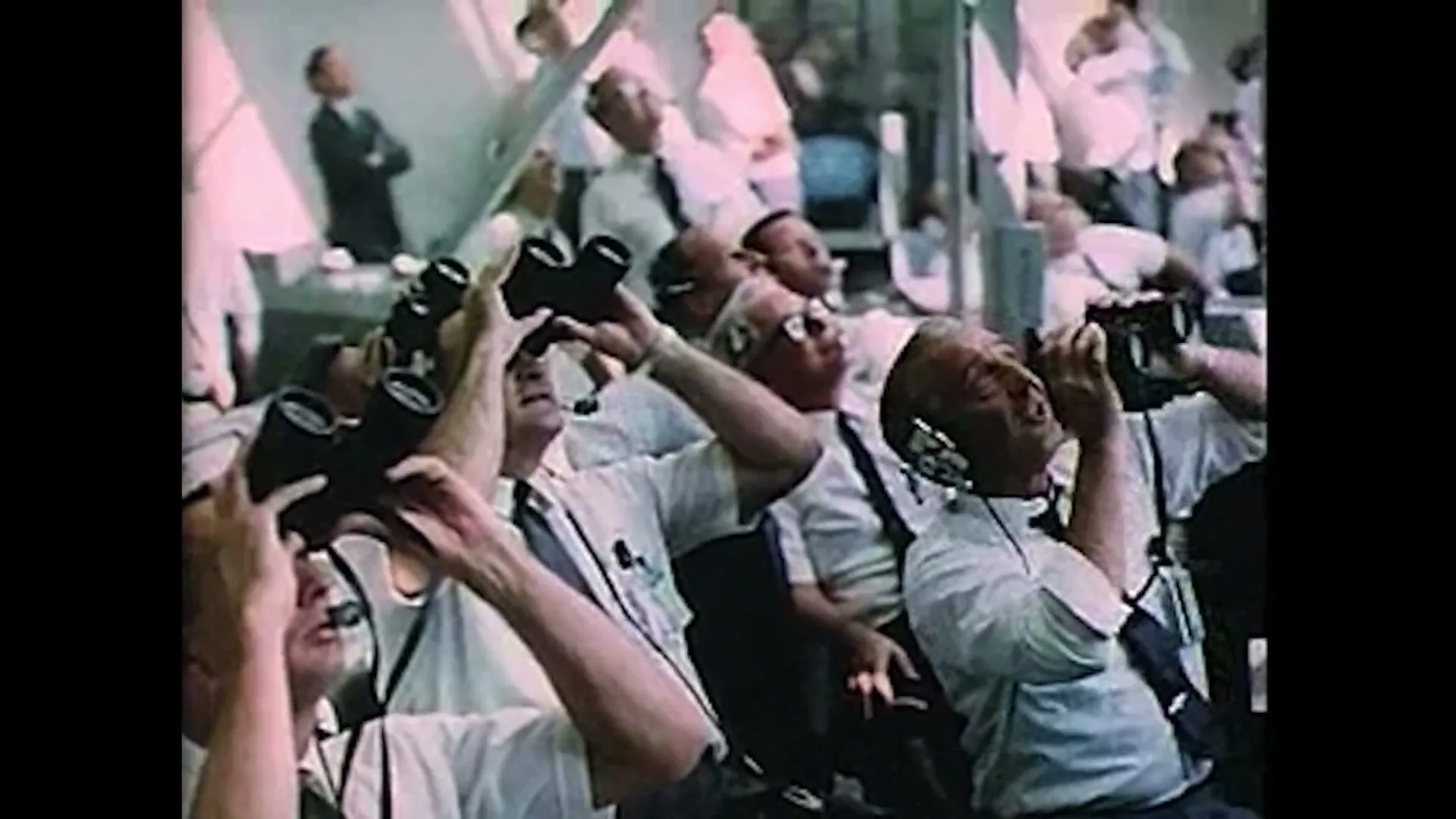 A group of male scientists and engineers in white shirts and ties look up, some with binoculars, in a control room.