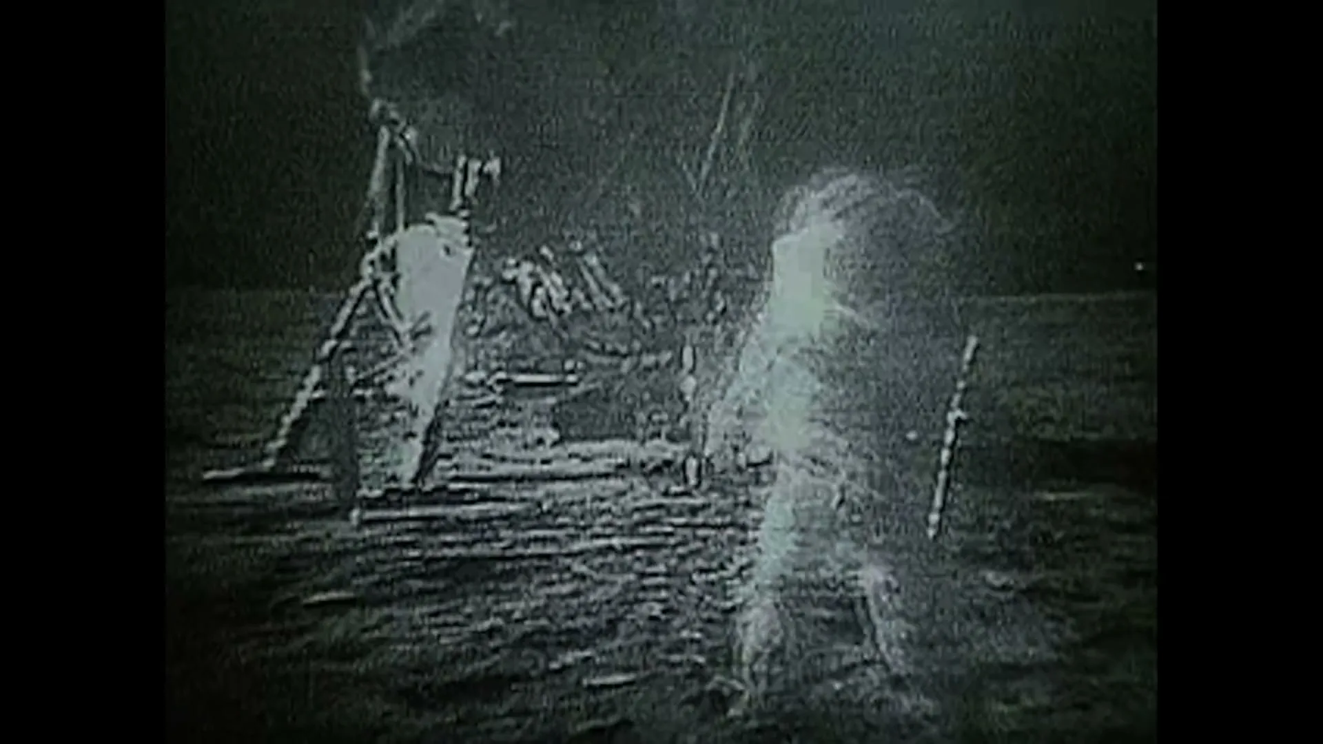 A grainy, black and white archival footage shot shows an astronaut standing next to a lunar lander on the moon's surface.