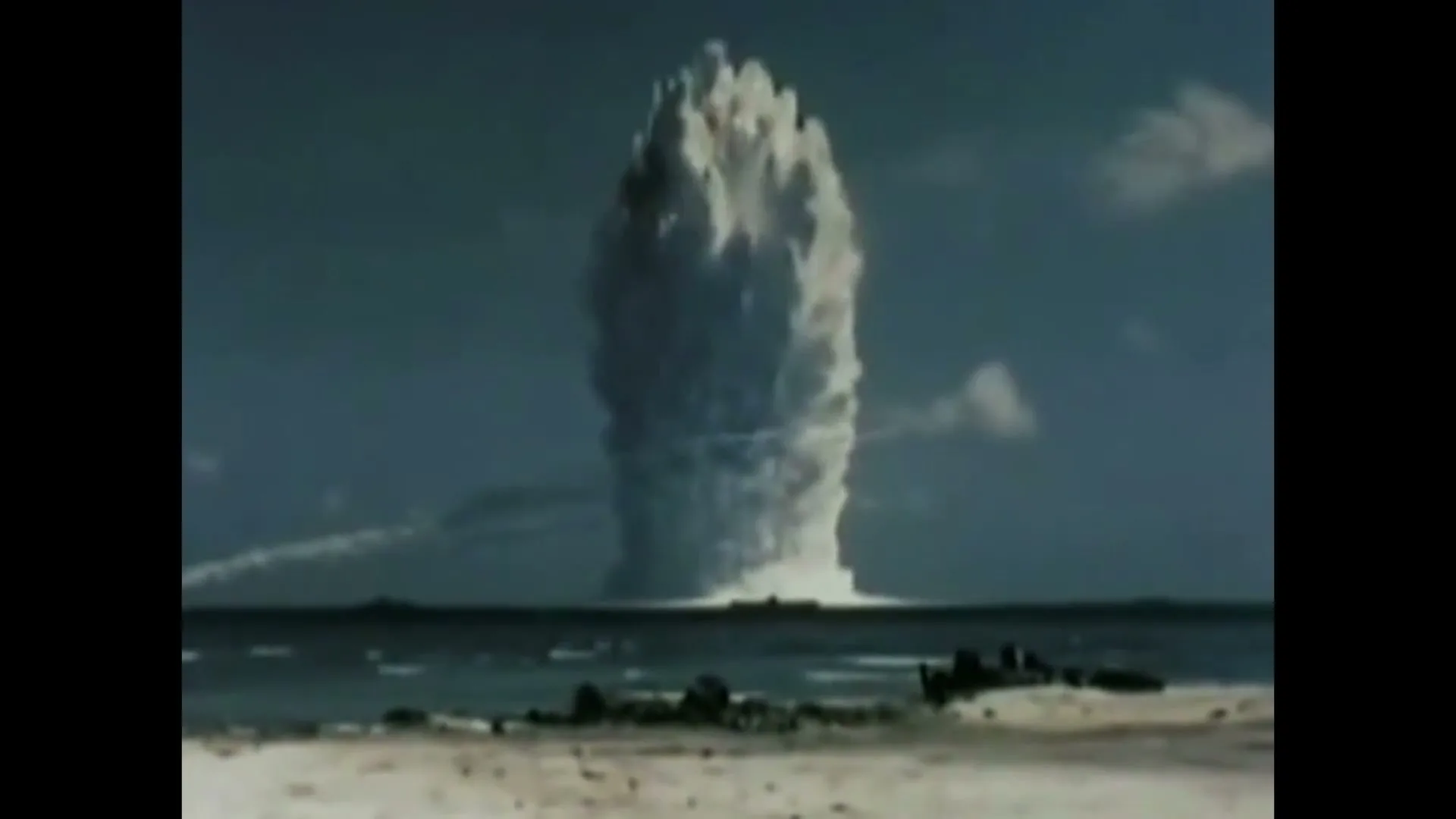 Archival footage shows a massive explosion erupting from the ocean, sending a huge column of water and smoke into the sky near a sandy shore.