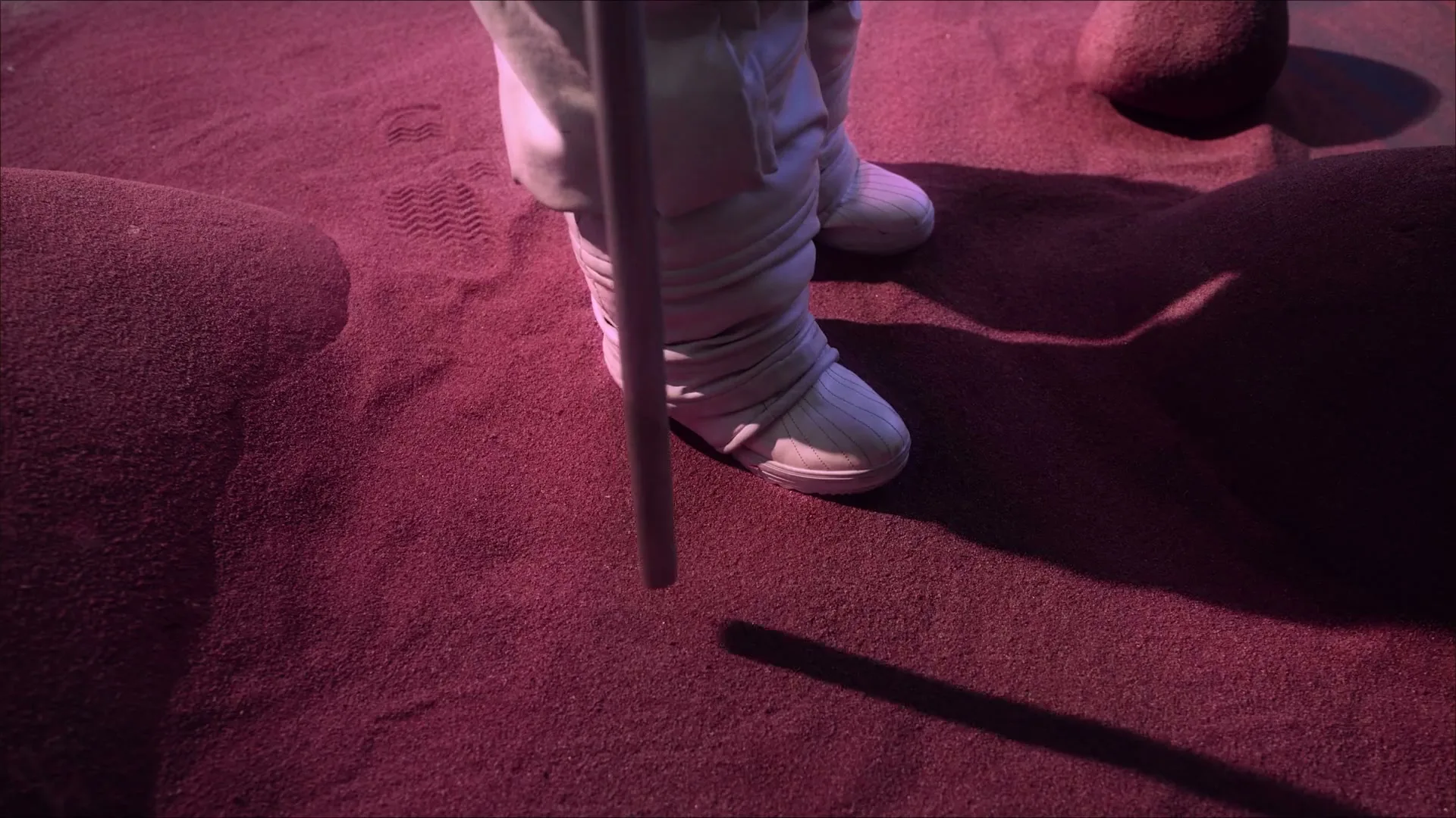 An astronaut's feet in white boots stand on a red, sandy, alien-like terrain, holding a staff that casts a long shadow.