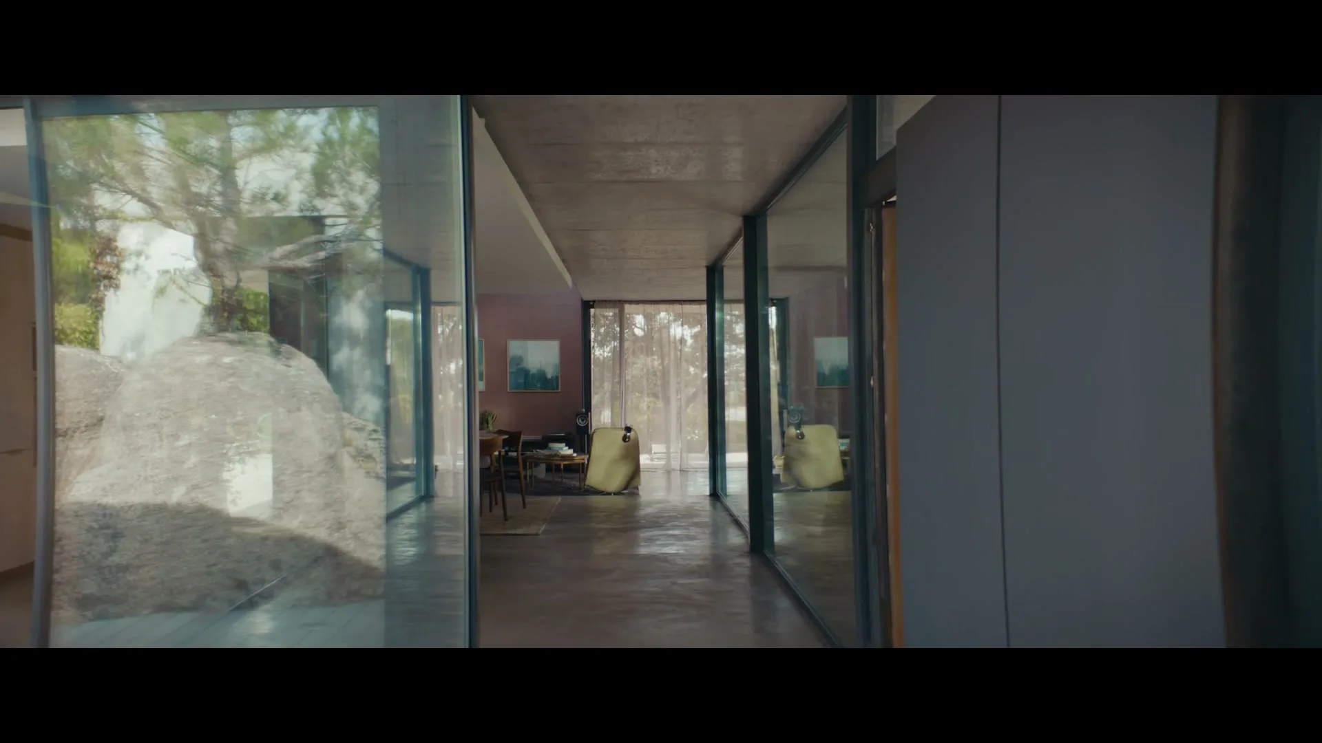 A man in a grey jacket and sunglasses walks into a modern house with glass walls, carrying a paper bag.
