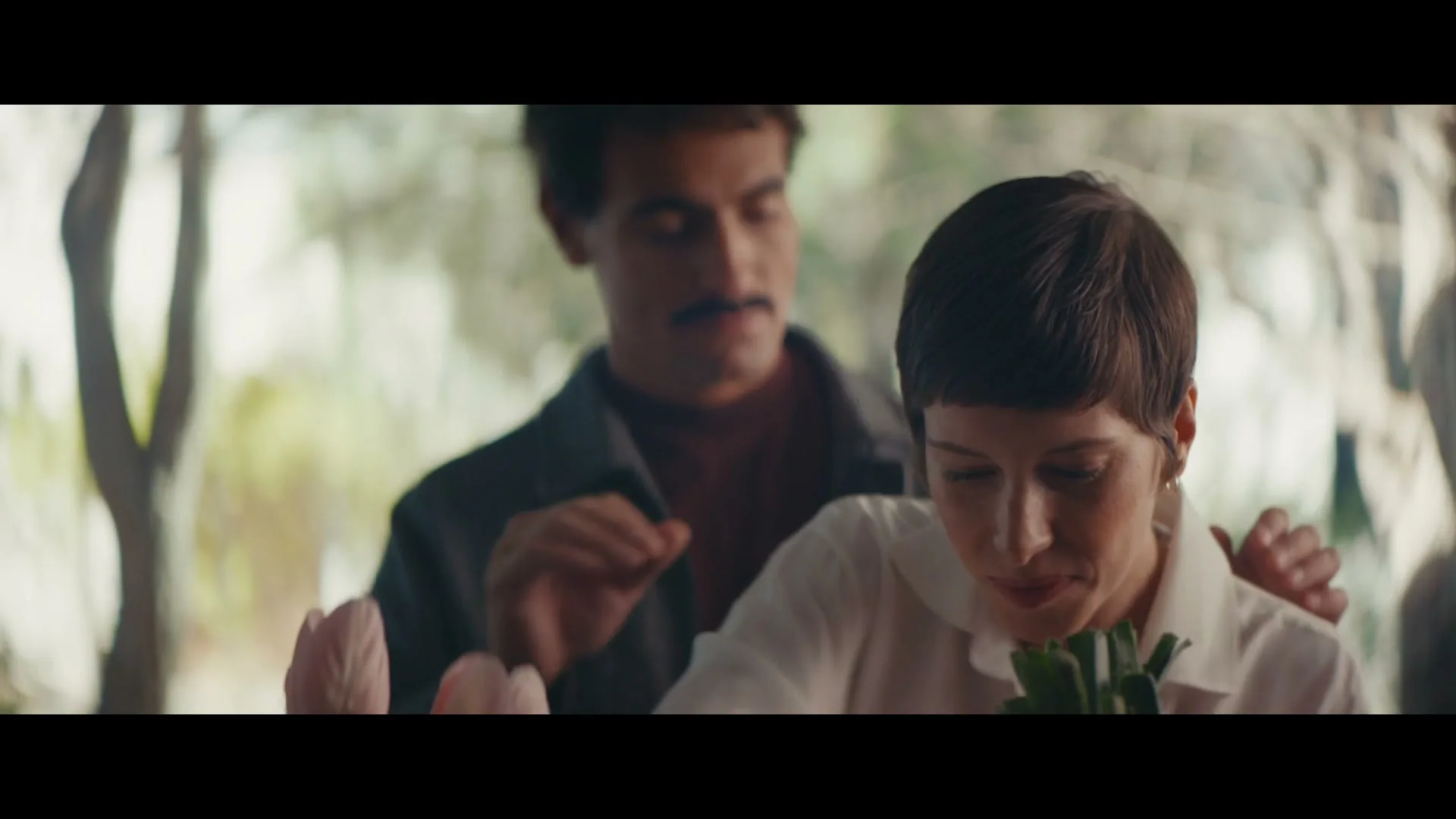 A woman with short hair smiles as a man with a mustache stands behind her, gently touching her shoulders in a bright, outdoor setting.