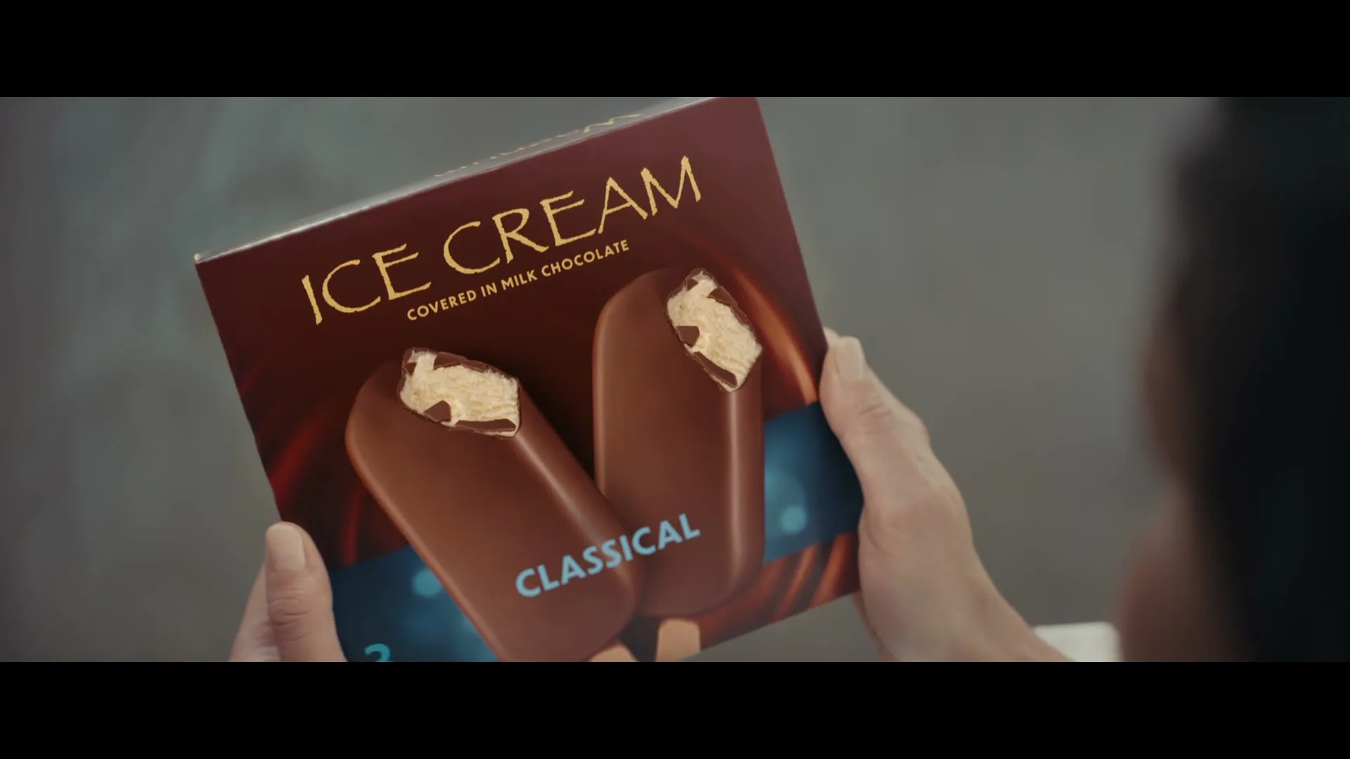 A person's hands hold a box of "ICE CREAM CLASSICAL" with two chocolate-covered ice cream bars on the front, in an interior setting.