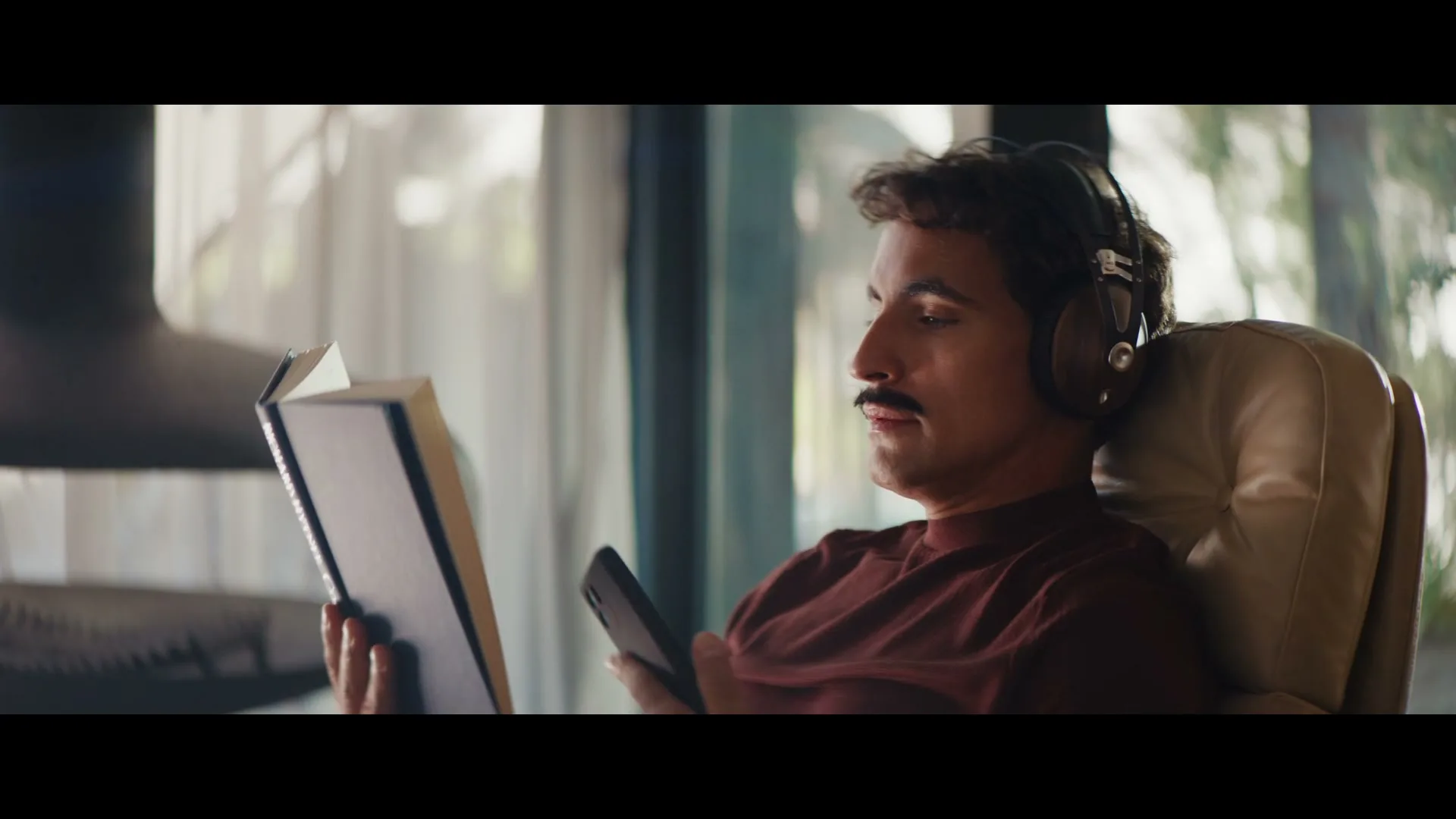 A man with a mustache and headphones sits in a chair, reading a book and looking at his phone in a sunlit room.