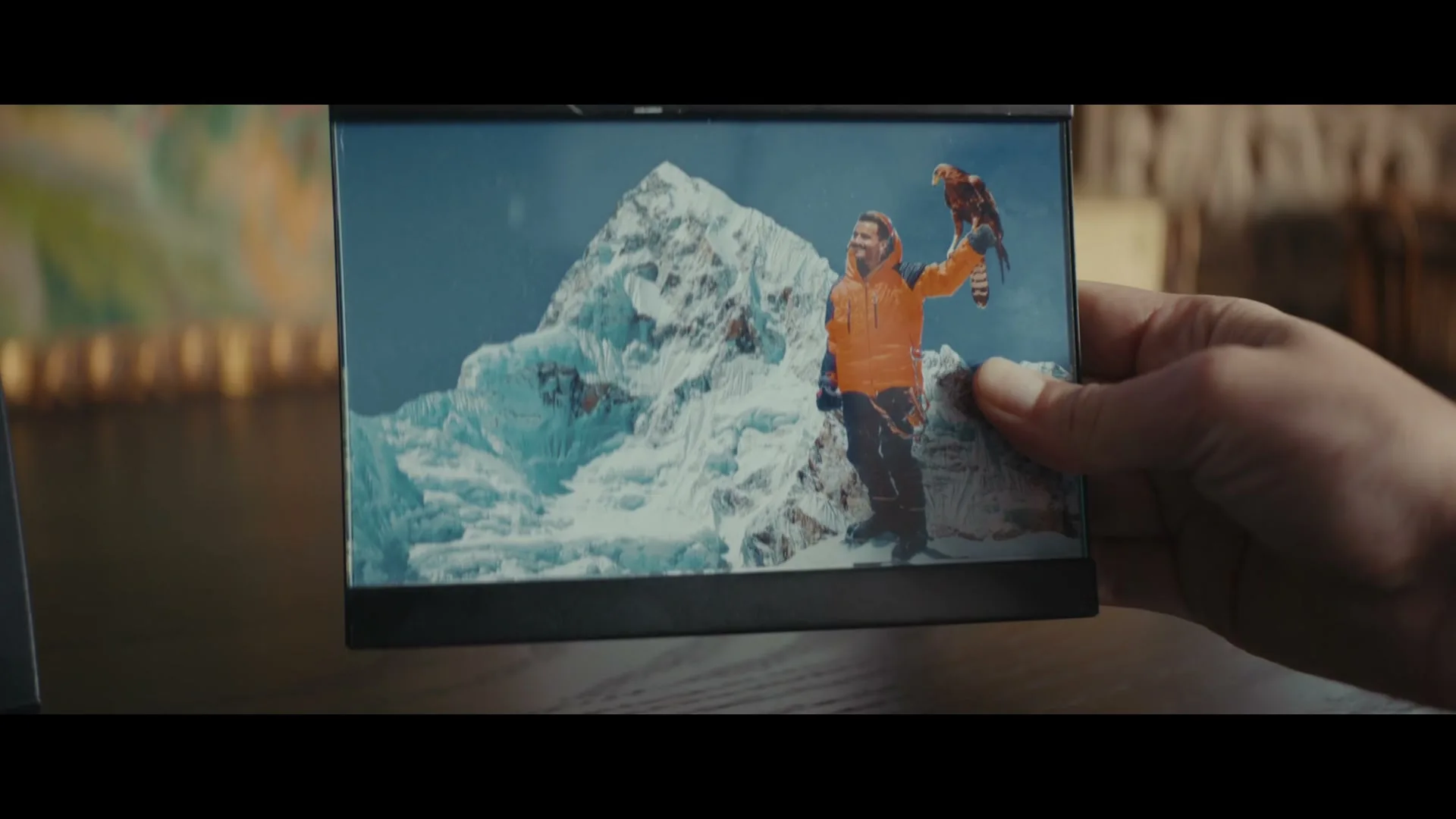 A hand holds a small digital photo frame displaying a man in an orange jacket with an eagle on his arm, standing on a snowy mountain.