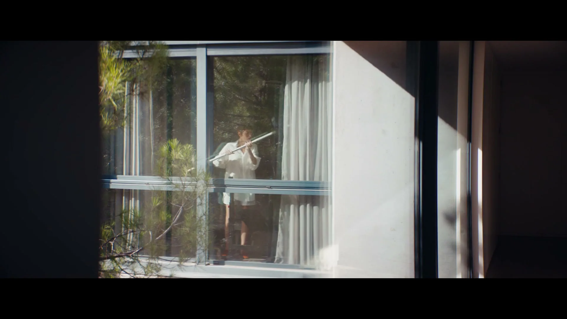 A young woman in a white shirt and shorts is seen through a window, playing an air guitar with a broomstick in a modern house.