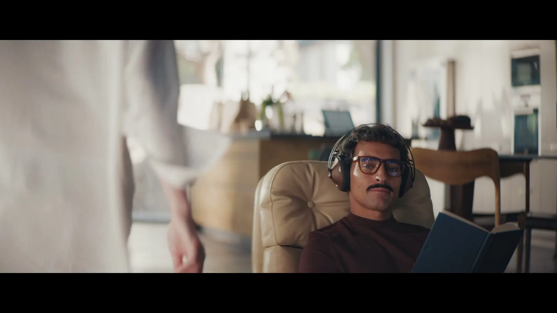 A man with a mustache and glasses, wearing headphones, sits in a chair reading a book while a person walks past him in a modern living room.