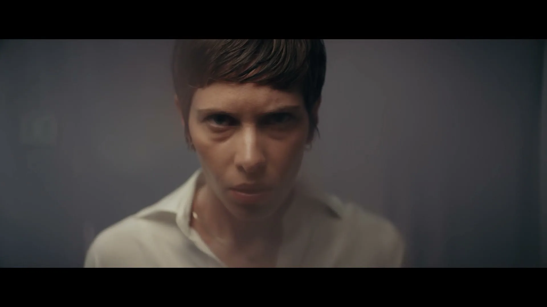 A woman with short brown hair and a white shirt stares intensely at the camera in a dimly lit room.