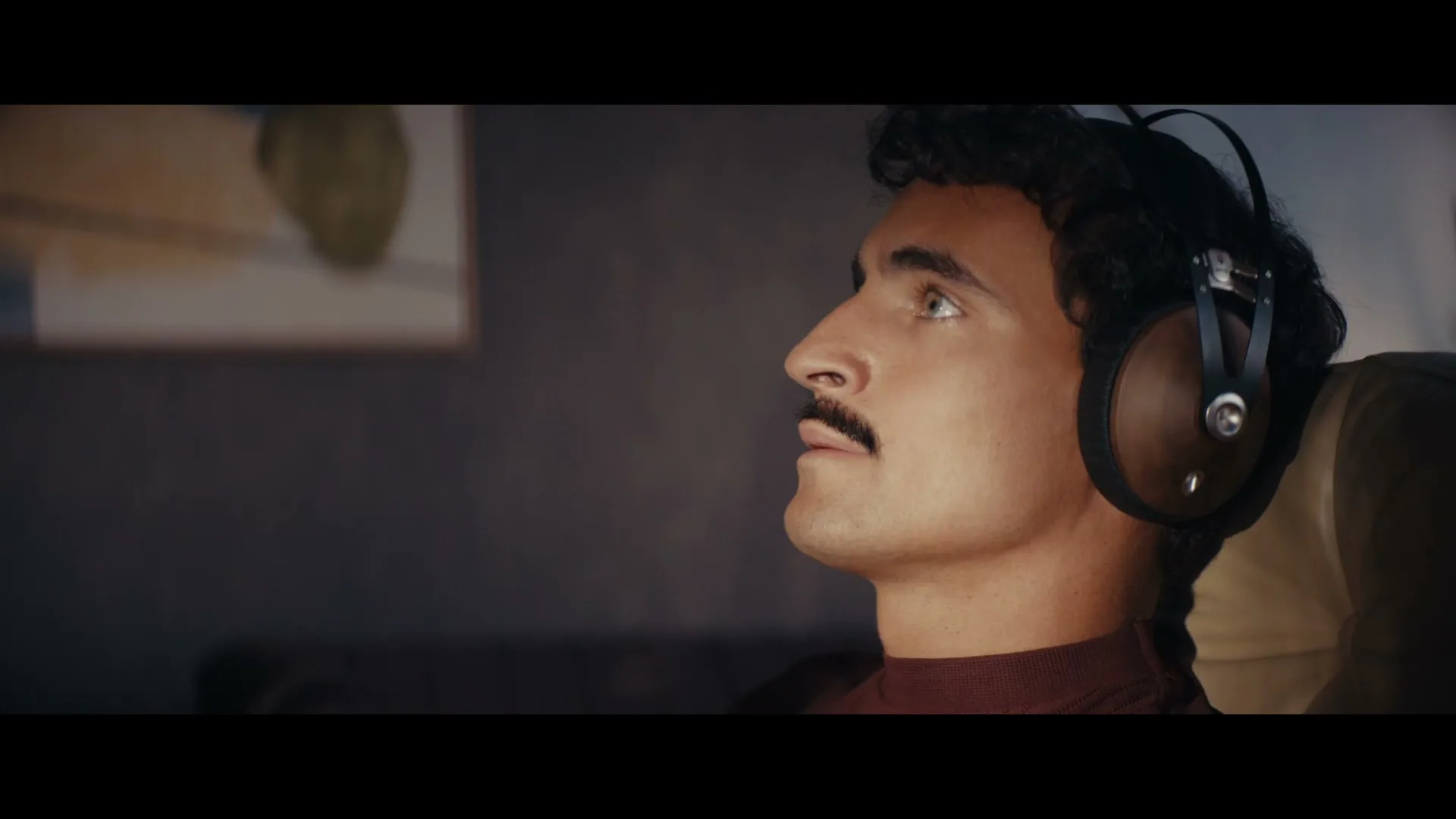 A man with a mustache and headphones looks up with a slightly open mouth, in a dimly lit room.