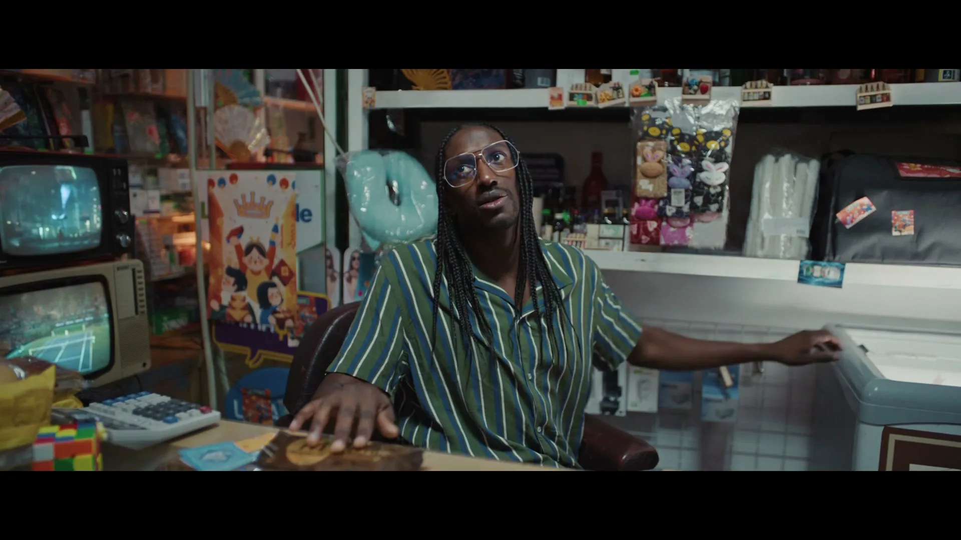 A man with dreadlocks and glasses, wearing a striped shirt, sits behind a counter in a cluttered convenience store, looking up at someone off-screen.
