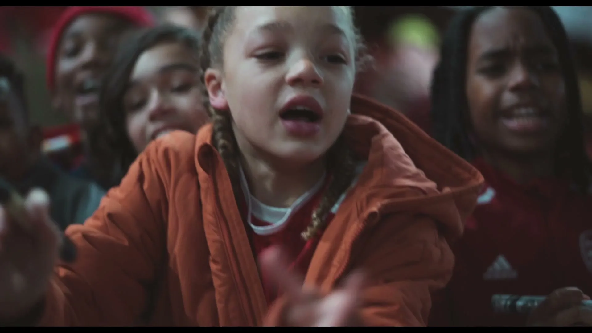 A young girl with braided hair, wearing an orange jacket, shouts excitedly in a crowd of children at a sports event.