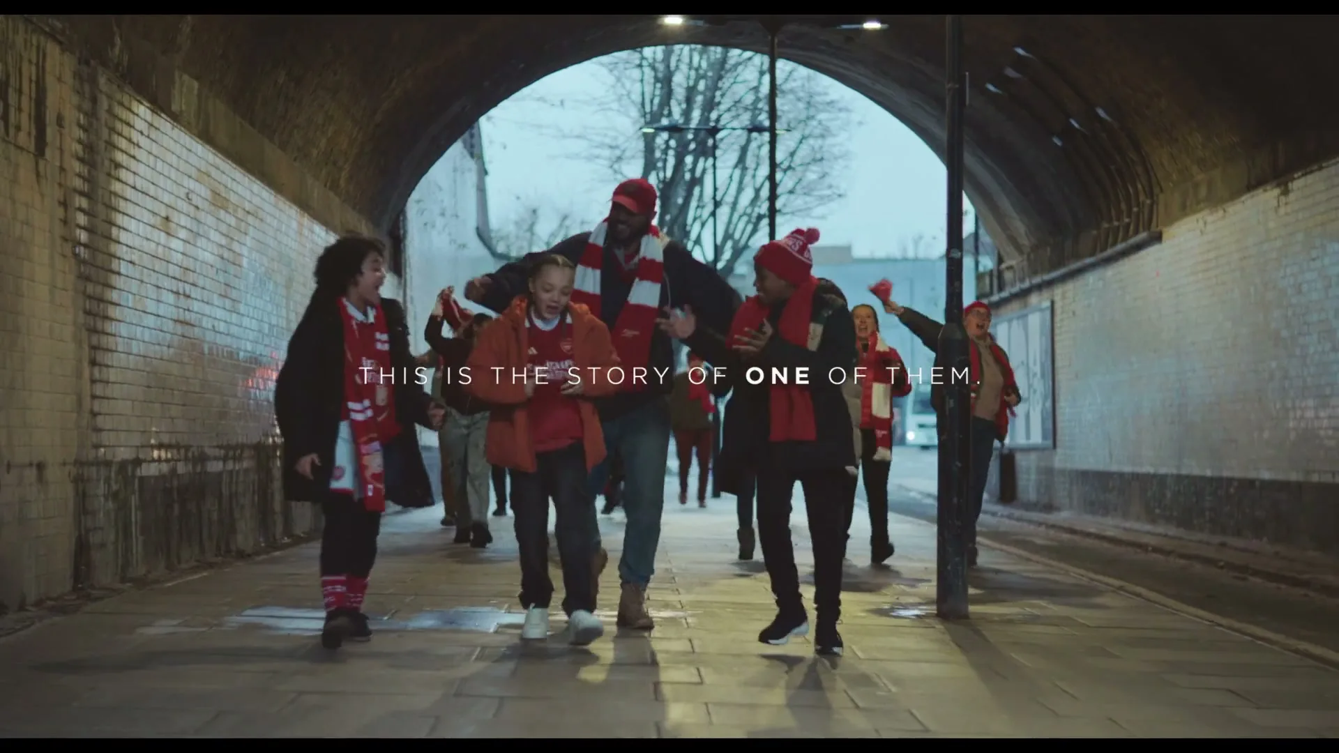 A diverse group of football fans, including children and adults, walk together through a tunnel, wearing red and white team merchandise.