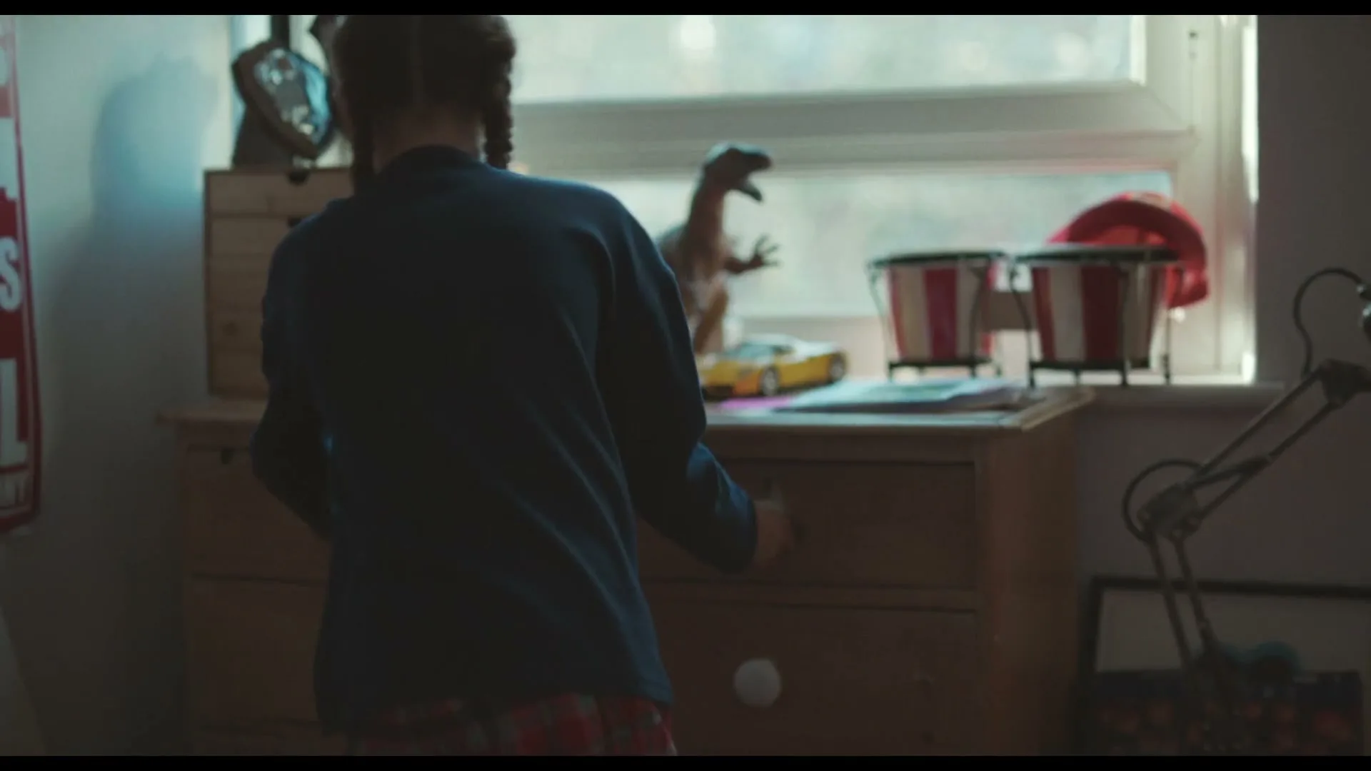 A young girl with braided hair, wearing a blue top and plaid pajamas, stands with her back to the camera, opening a drawer in a wooden dresser in her bedroom.