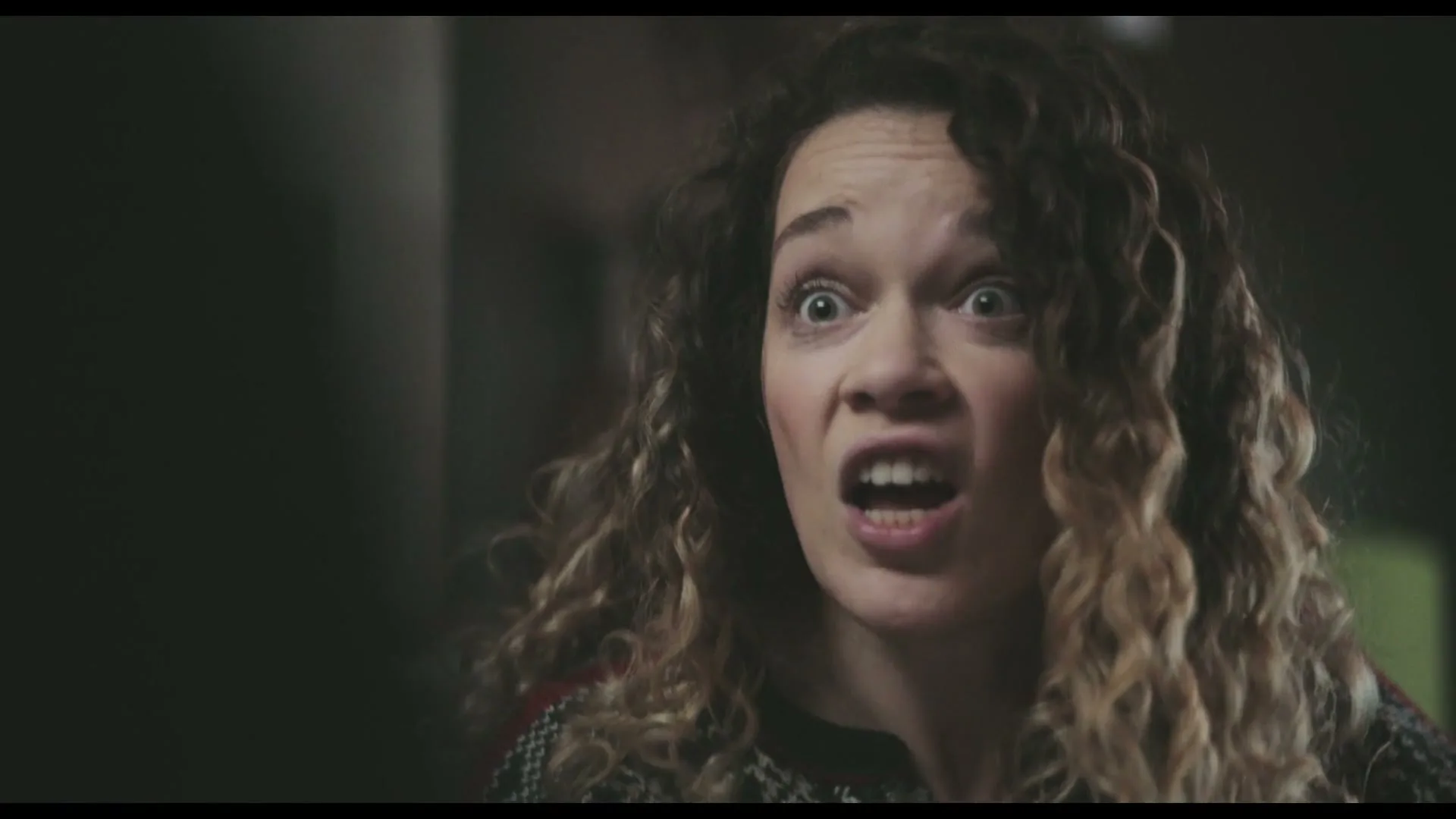 A woman with curly hair and a patterned sweater looks shocked and surprised, with wide eyes and an open mouth, in an interior setting.