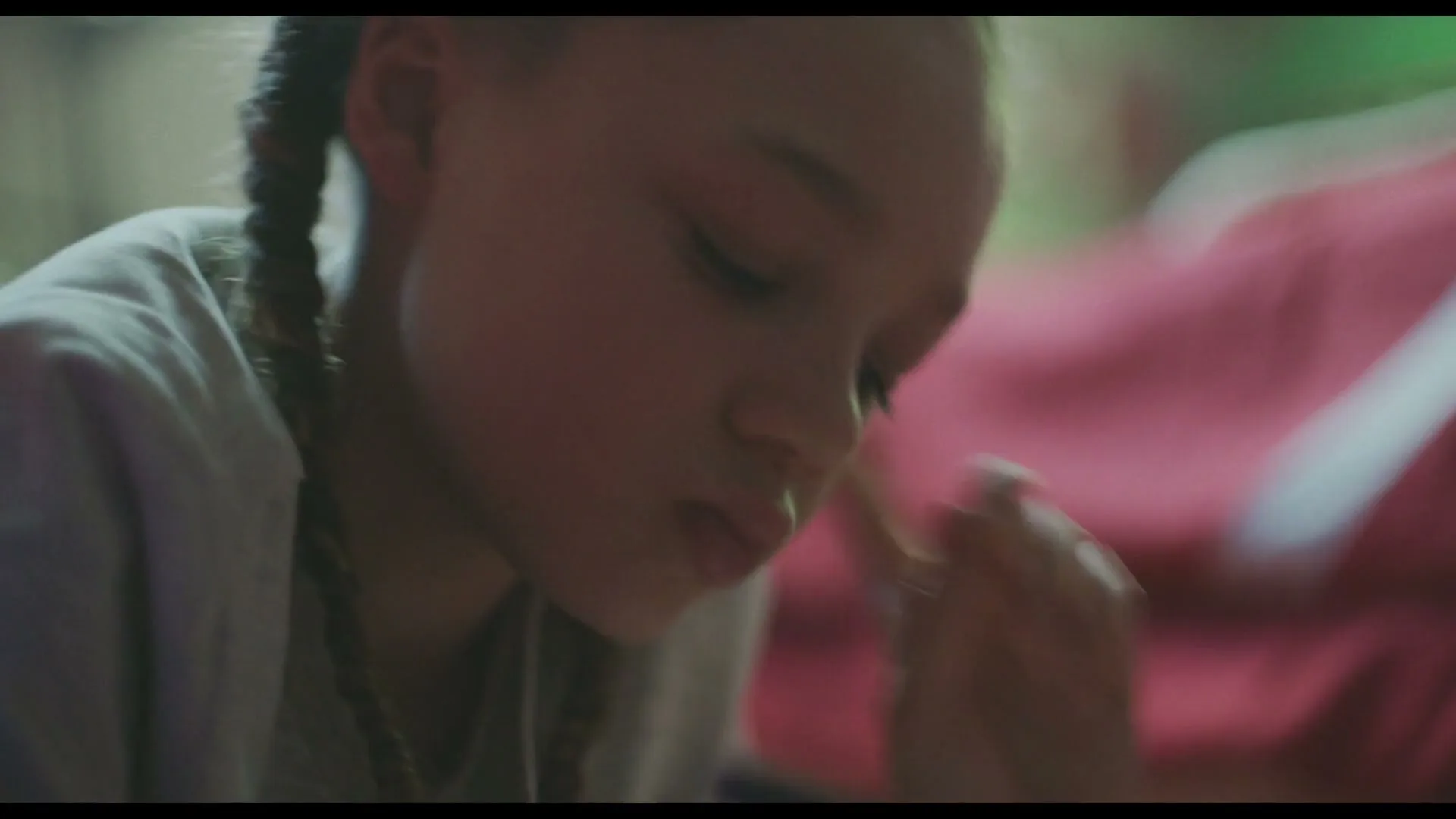A young girl with braided hair, wearing a light-colored top, looks down intently while drawing or writing, with a red object blurred in the background.