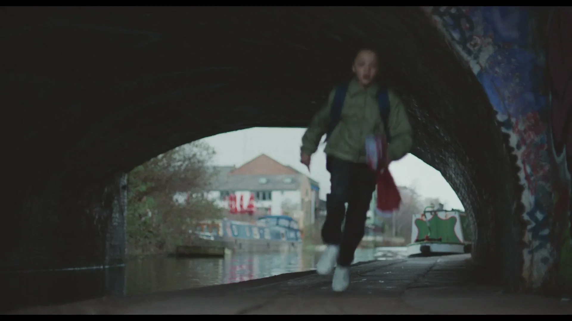 A young boy, wearing a green jacket and backpack, runs along a canal path under a bridge, holding a red item.