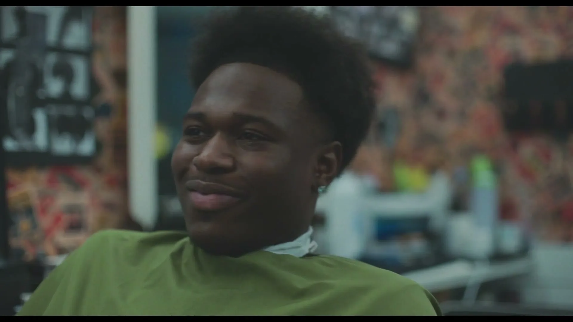 A young Black man with a fresh haircut and a green barber's cape smiles and laughs while sitting in a barber shop.