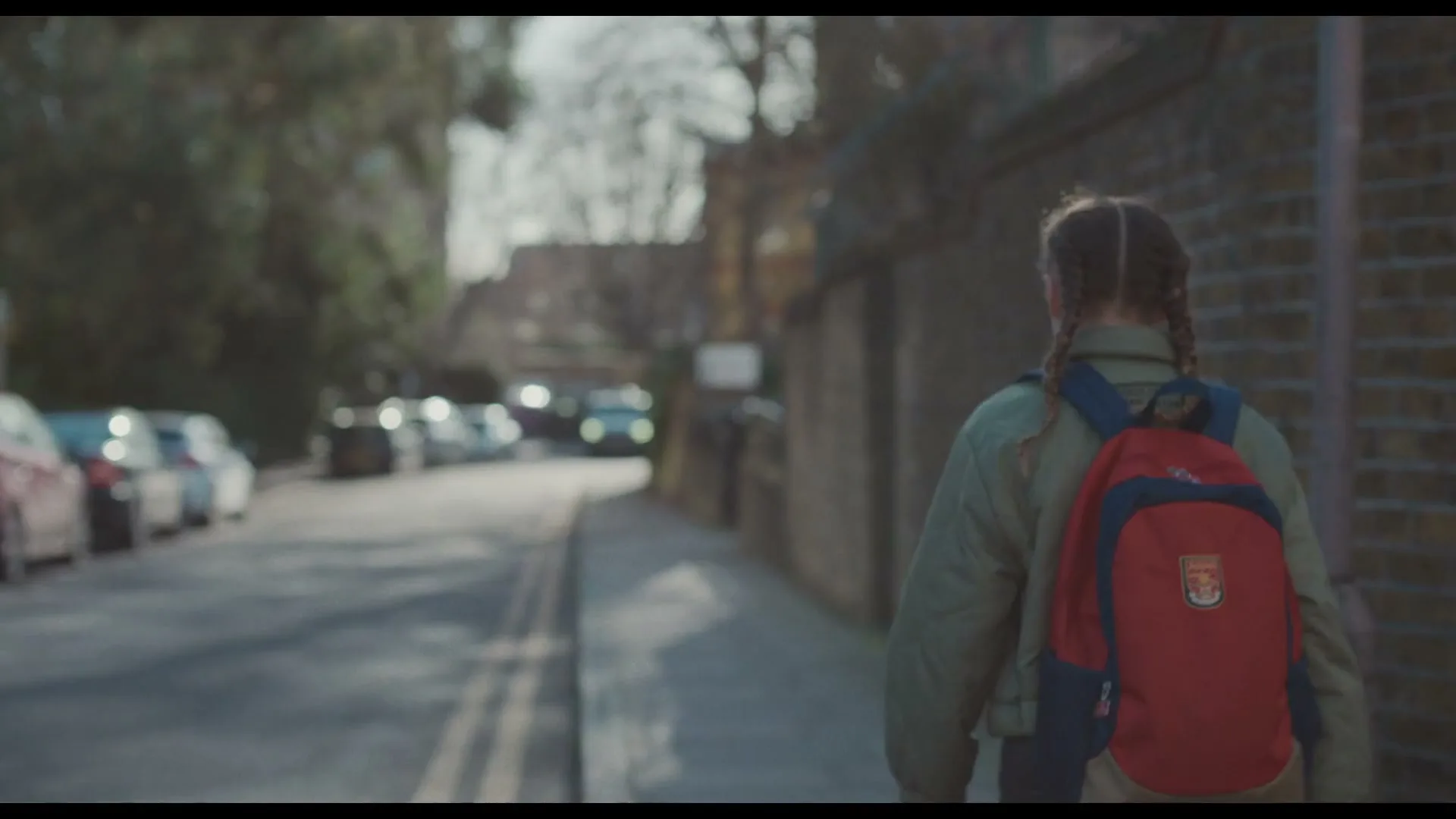 A young girl with braided hair and a red backpack walks away from the camera down a sunny street lined with parked cars and brick walls.