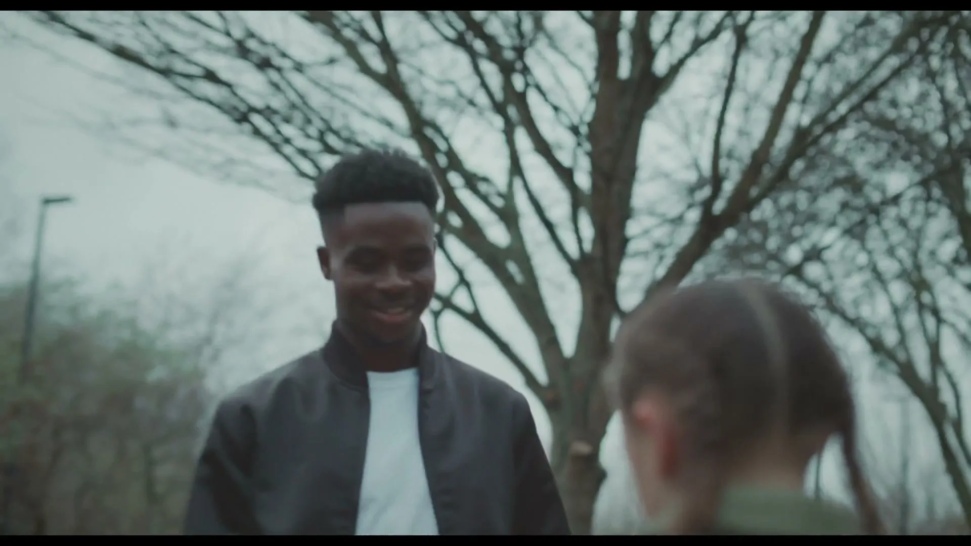 A young Black man in a black bomber jacket and white t-shirt smiles at a young girl with braided hair and a green jacket, who looks up at him with a hopeful expression on a cloudy day outdoors.