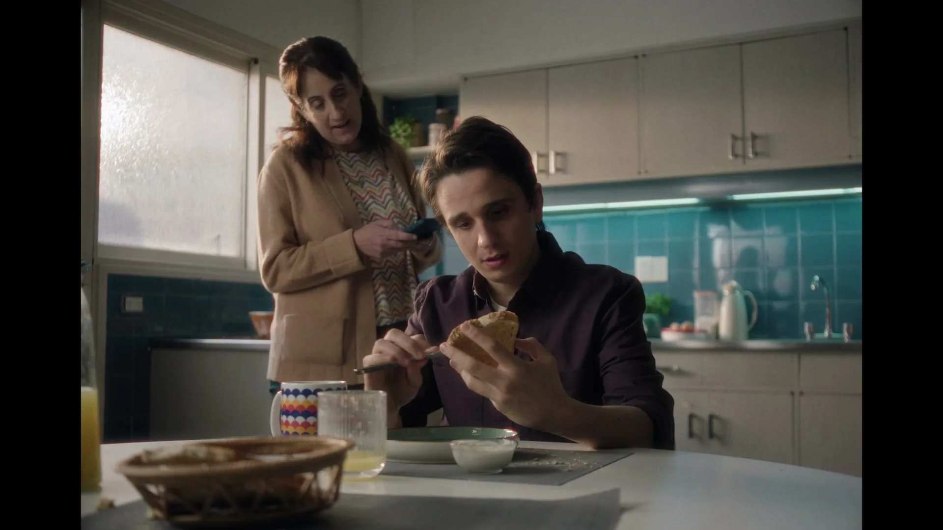 A young man sits at a kitchen table, spreading something on toast, while a woman stands behind him, looking at her phone.