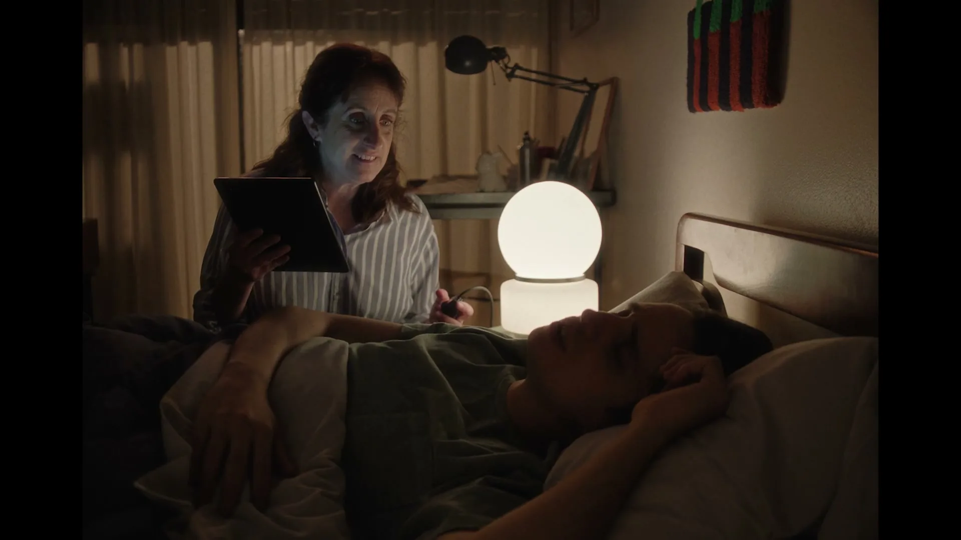 A mother sits on her bed, holding a tablet, looking at her son who is sleeping next to her.