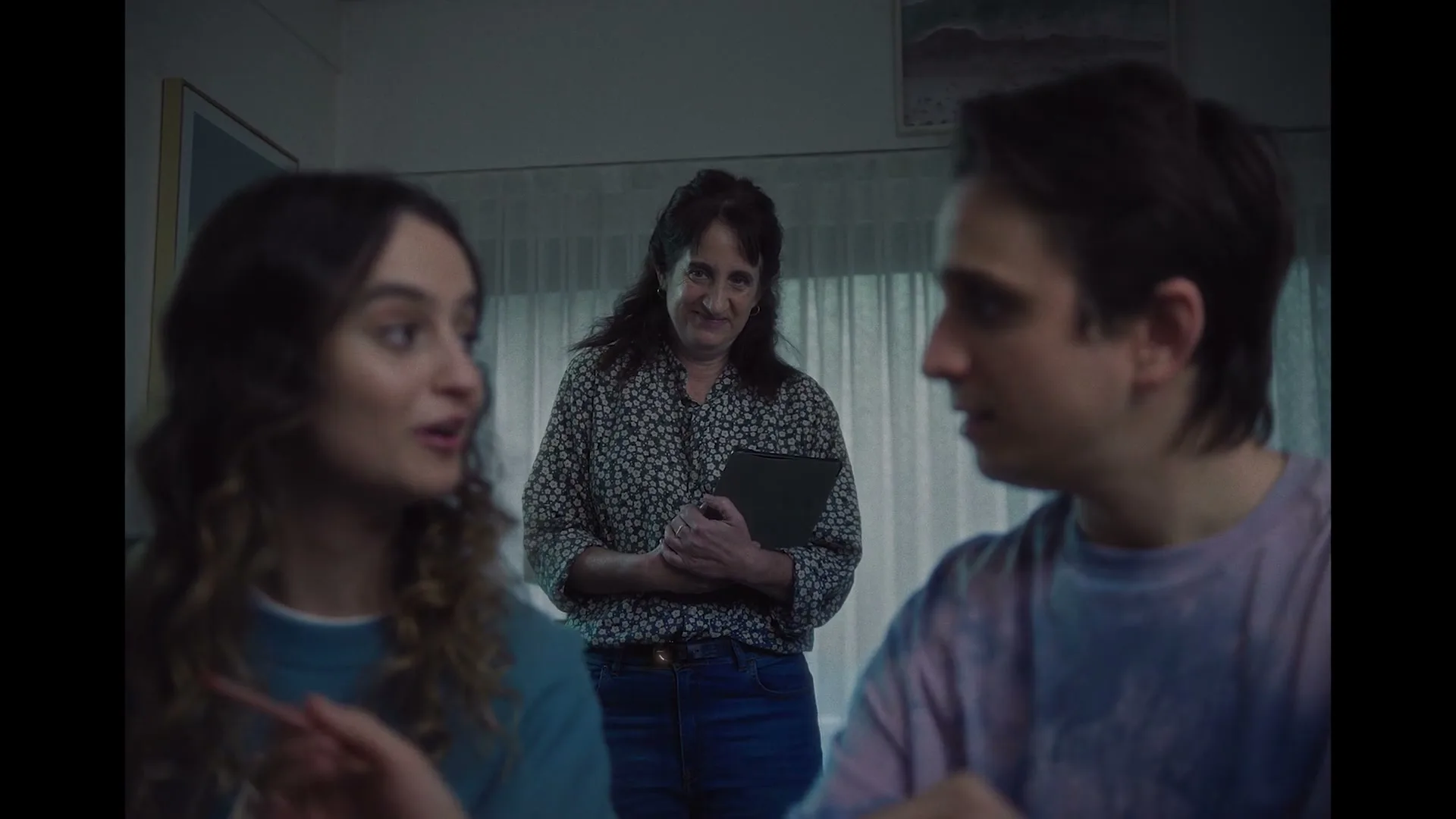 A mother stands in the background, holding a tablet, while two teenagers in the foreground talk and smile at each other in a dimly lit room.