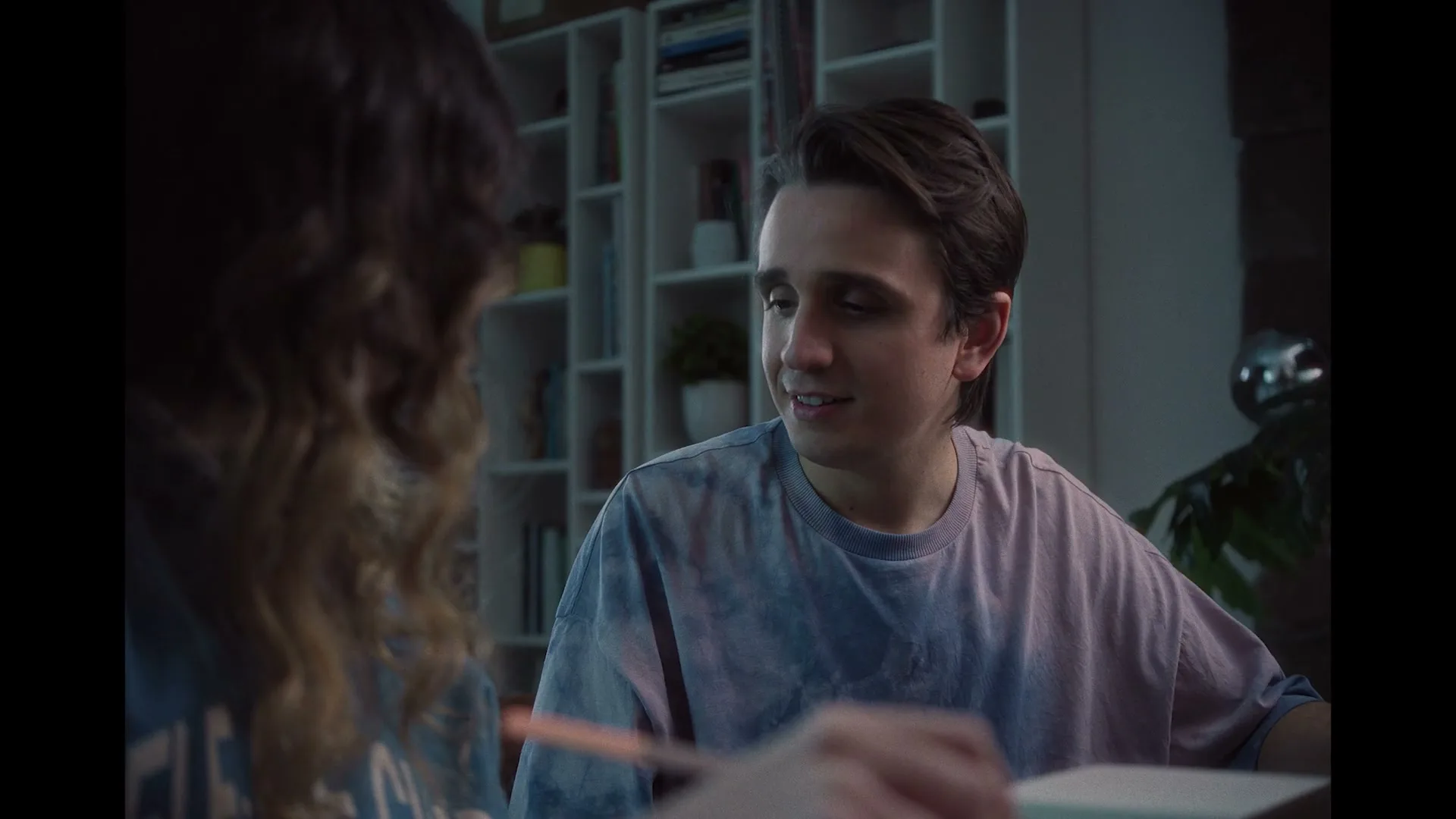 A young man with dark hair and a tie-dye shirt smiles at a young woman with curly hair, sitting at a table in a modern room with shelves.