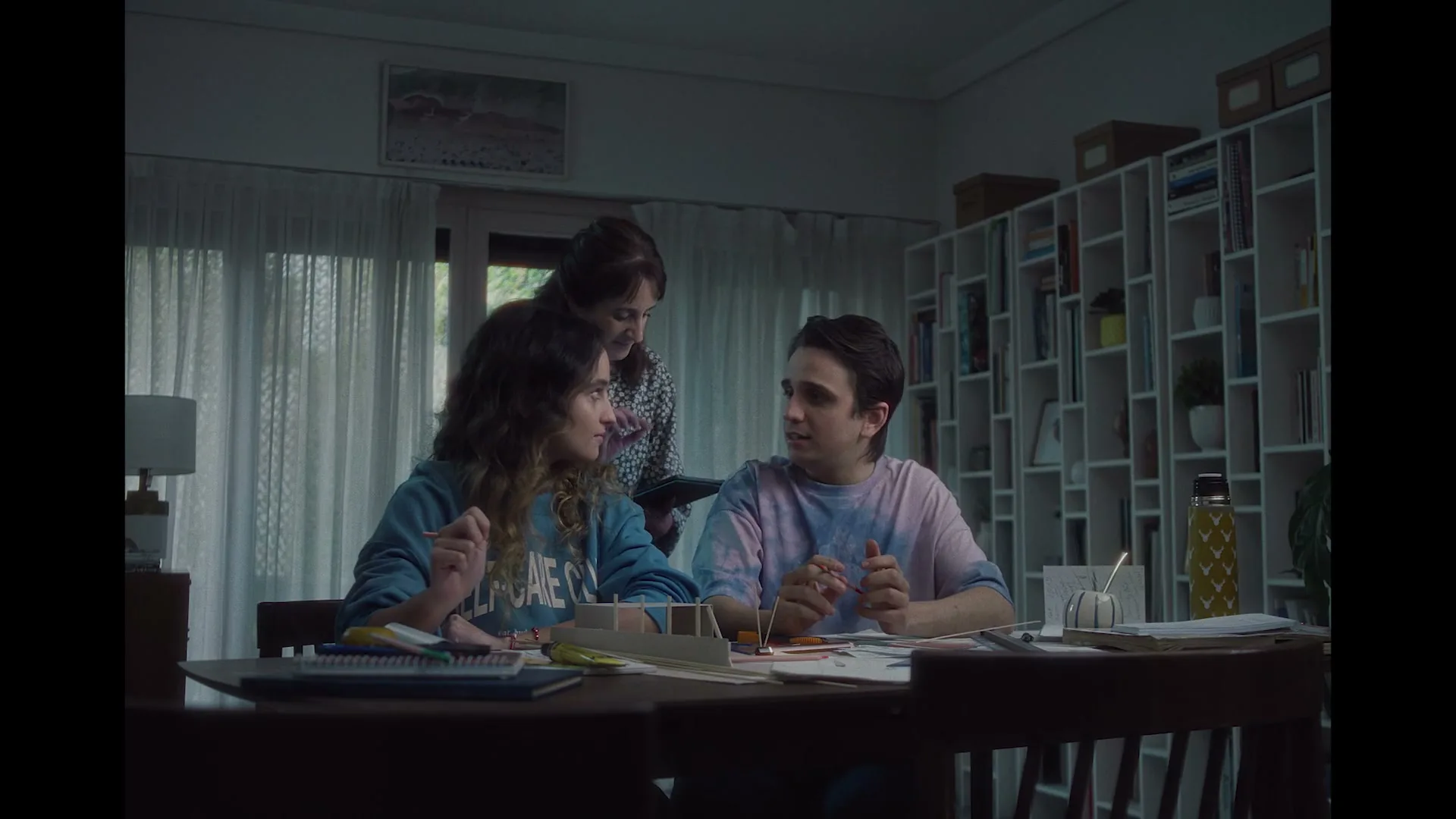 A mother approaches her two children, a girl and a boy, who are sitting at a dining table doing homework, in a dimly lit living room.