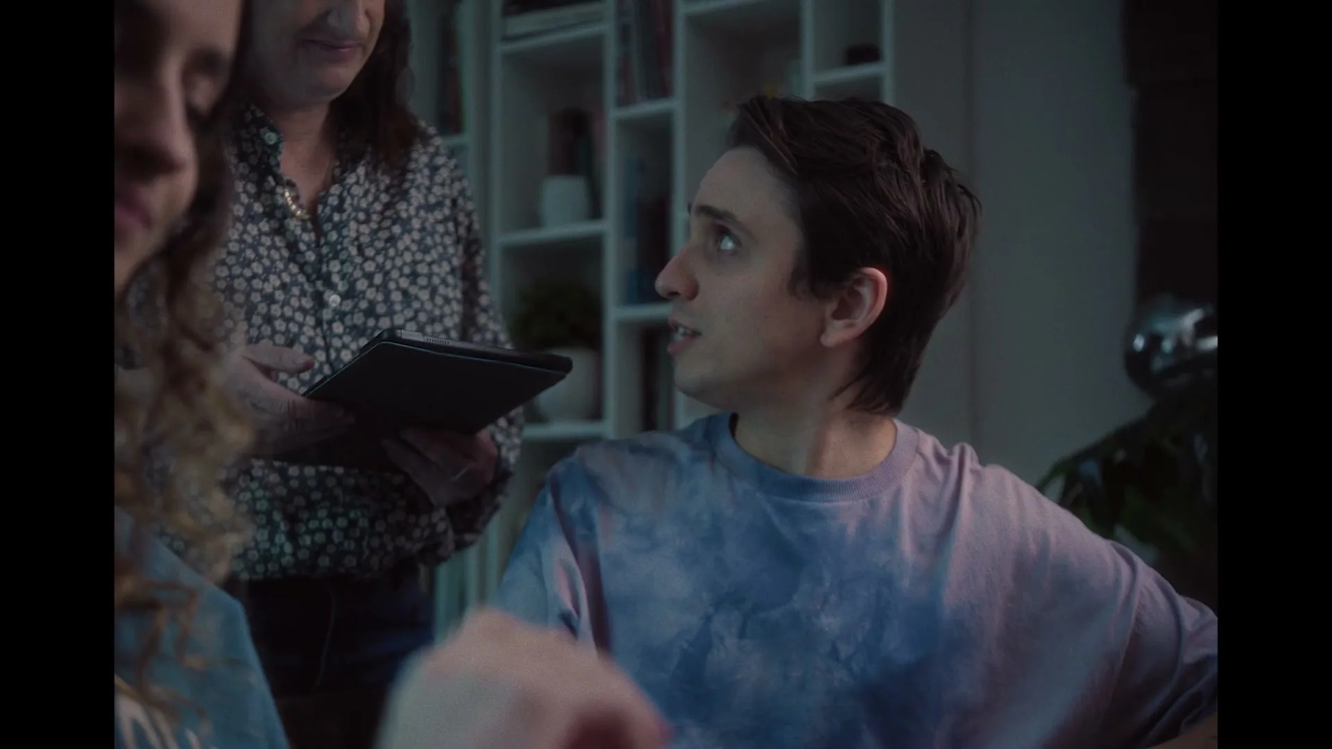 A young man in a tie-dye shirt looks up and to the right, while a woman holds a tablet behind him in a dimly lit room.