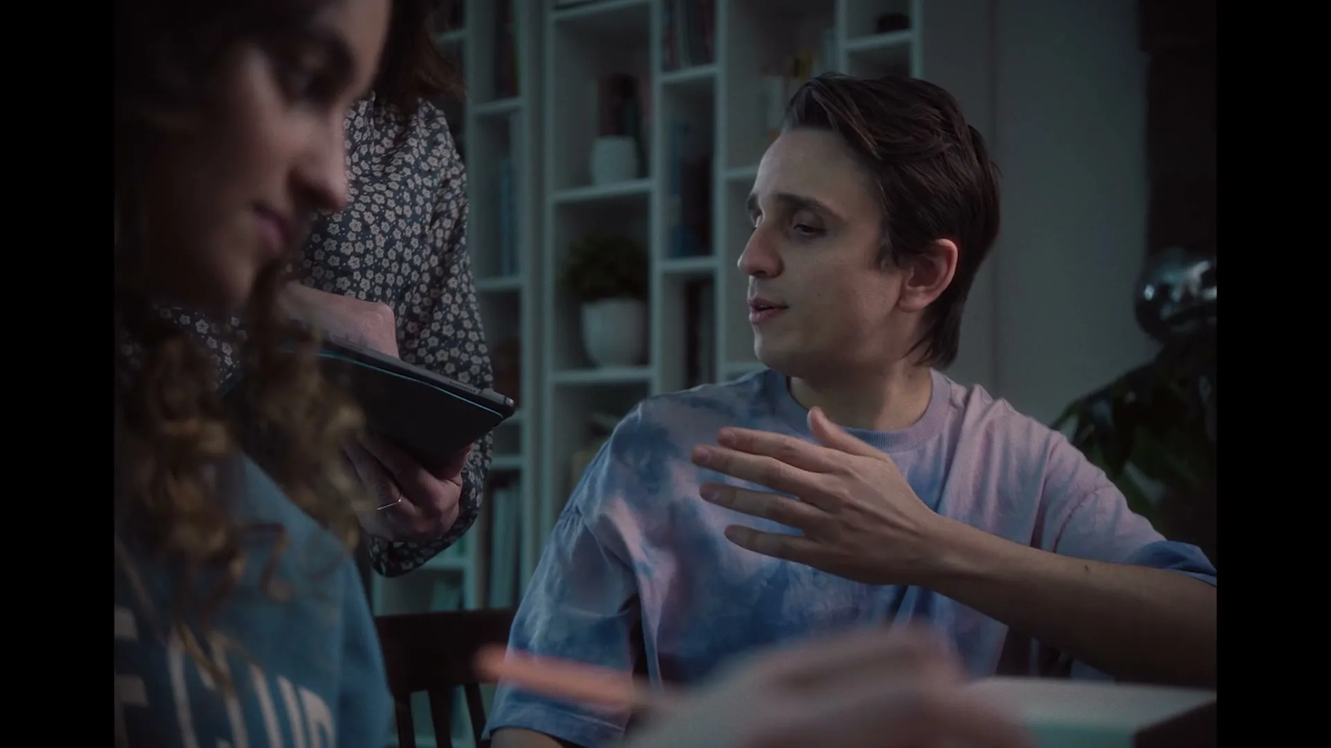 A young man in a tie-dye shirt talks to a young woman, who is partially visible, while another woman holds a tablet in the background, in an interior setting.