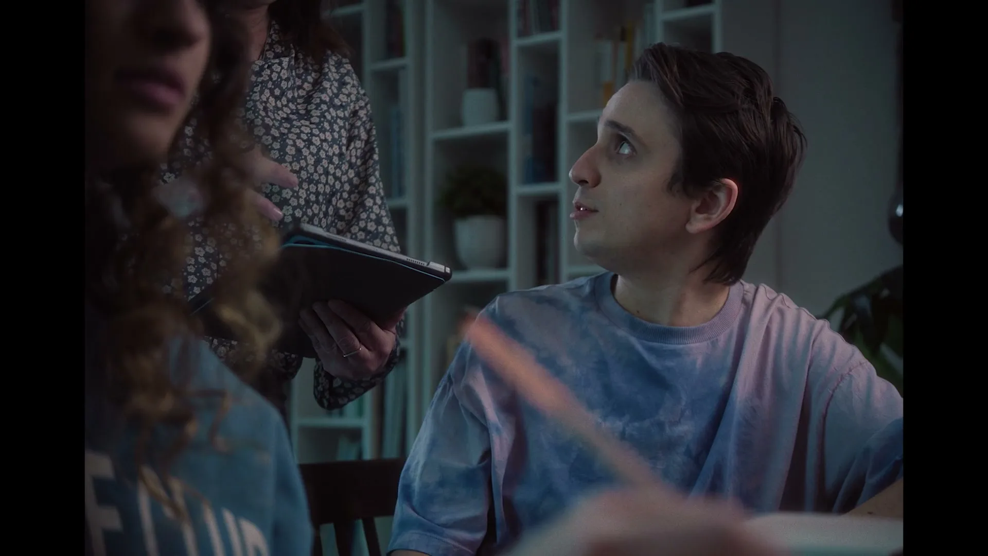 A young man in a tie-dye shirt looks up and to the right, while a woman in a floral shirt holds a tablet behind him in a dimly lit room.