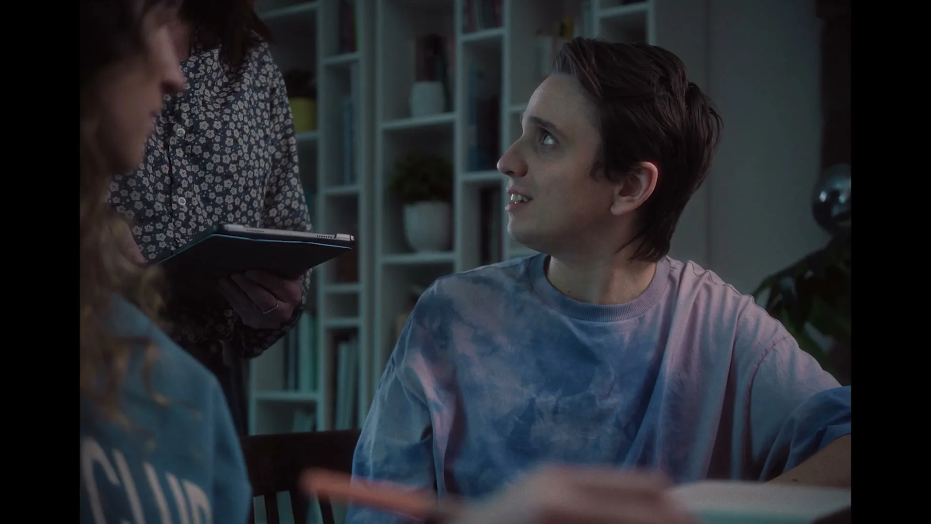 A young man in a tie-dye shirt talks to two women, one holding a tablet, in a modern living room at night.