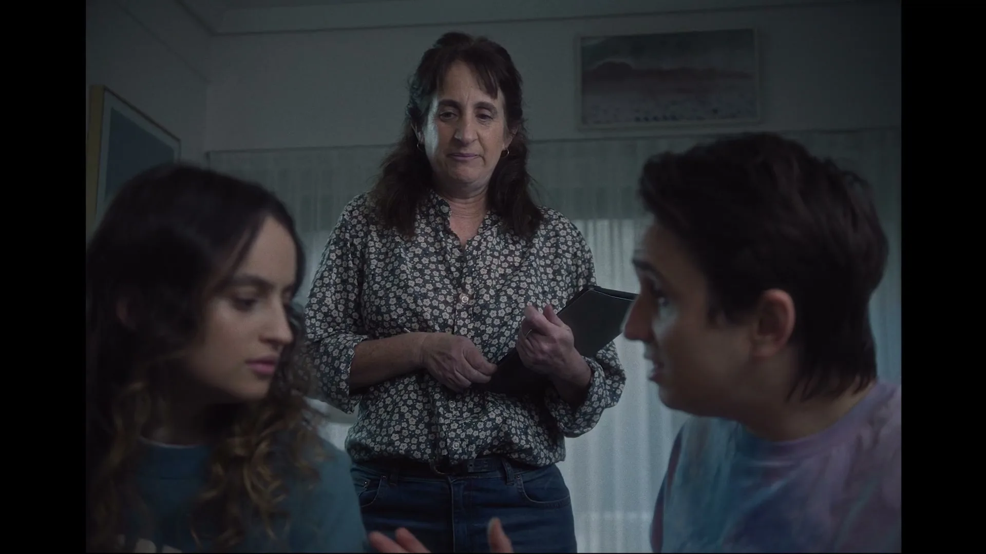 A mother stands between her two children, holding a tablet, as the children look at each other in a living room.