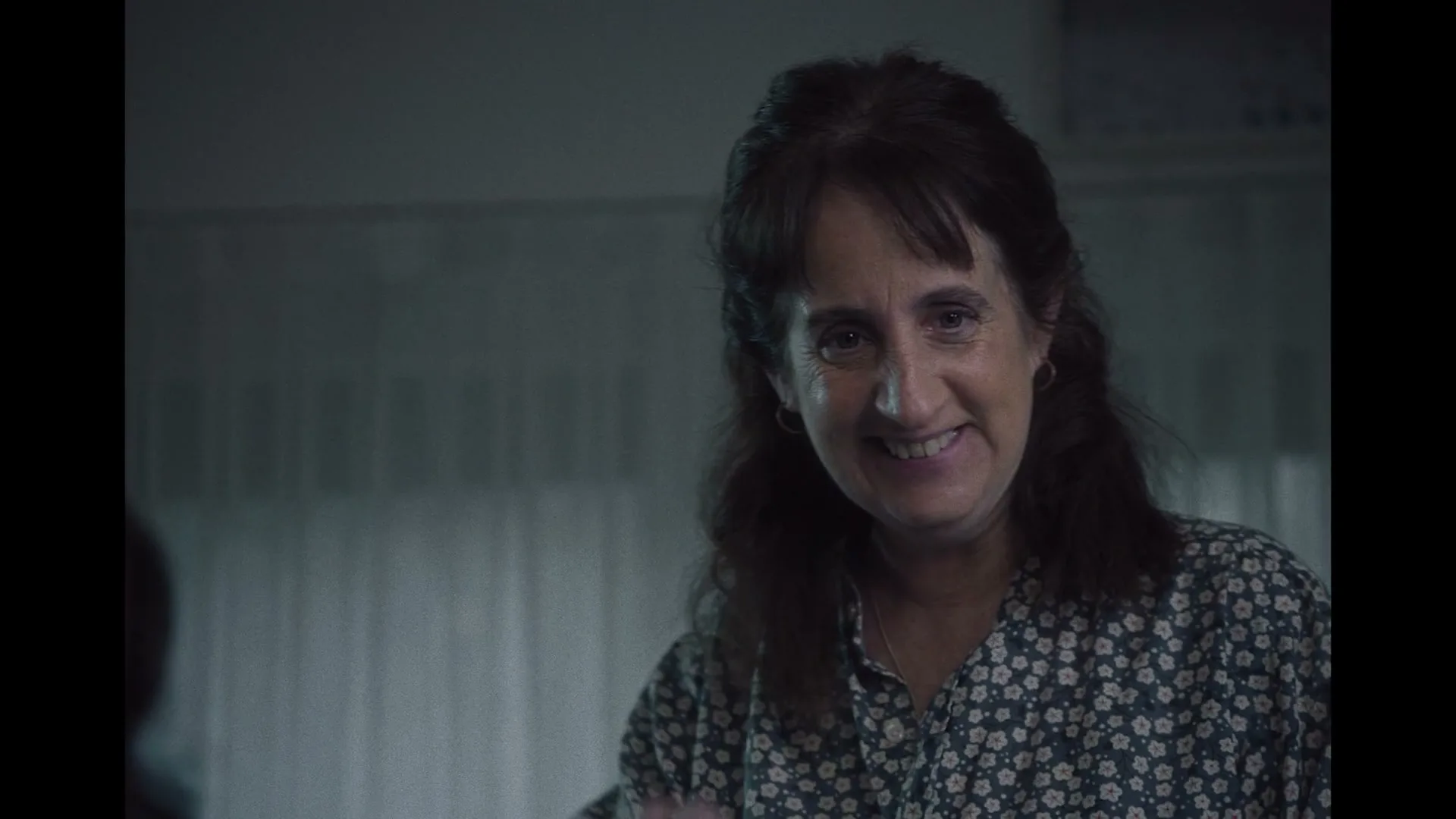 A middle-aged woman with dark hair and a floral shirt smiles, then turns away, in a dimly lit room.