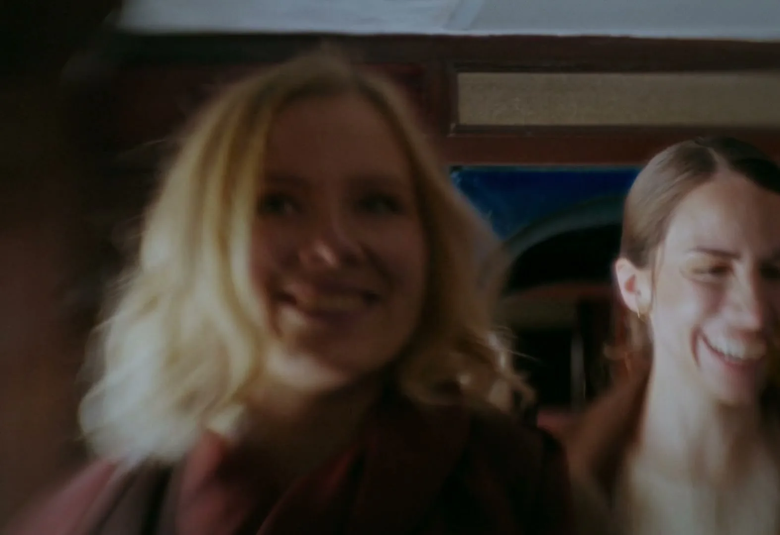 Two women, one with blonde hair and one with dark hair, laugh joyfully while walking through an interior space, possibly a train car.