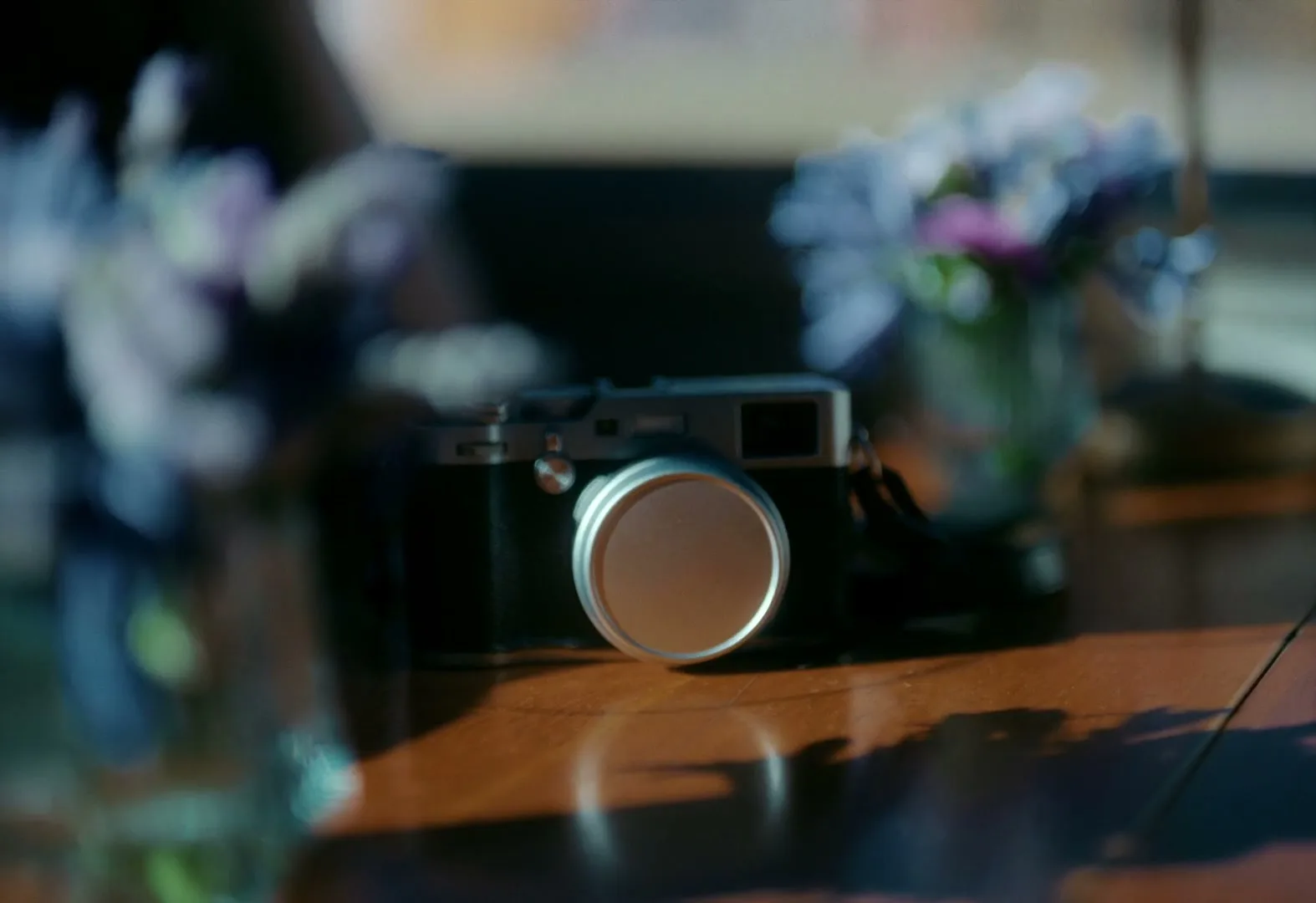 A vintage-style camera with a lens cap sits on a wooden table, with blurred flowers in the foreground and background, bathed in natural light.