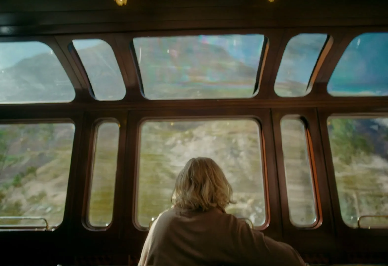 A blonde woman in a brown top looks out the window of a train at a mountainous landscape during the day.