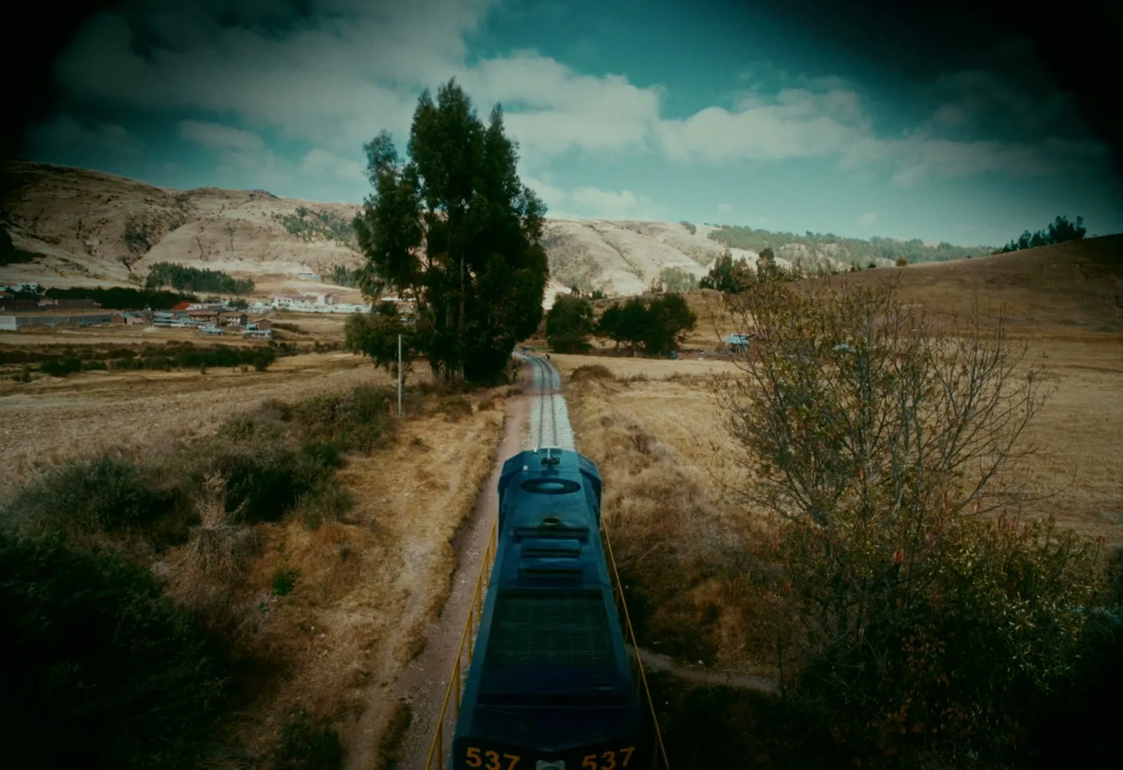 A train travels through a dry, rural landscape with hills and a small village in the distance under a cloudy sky.