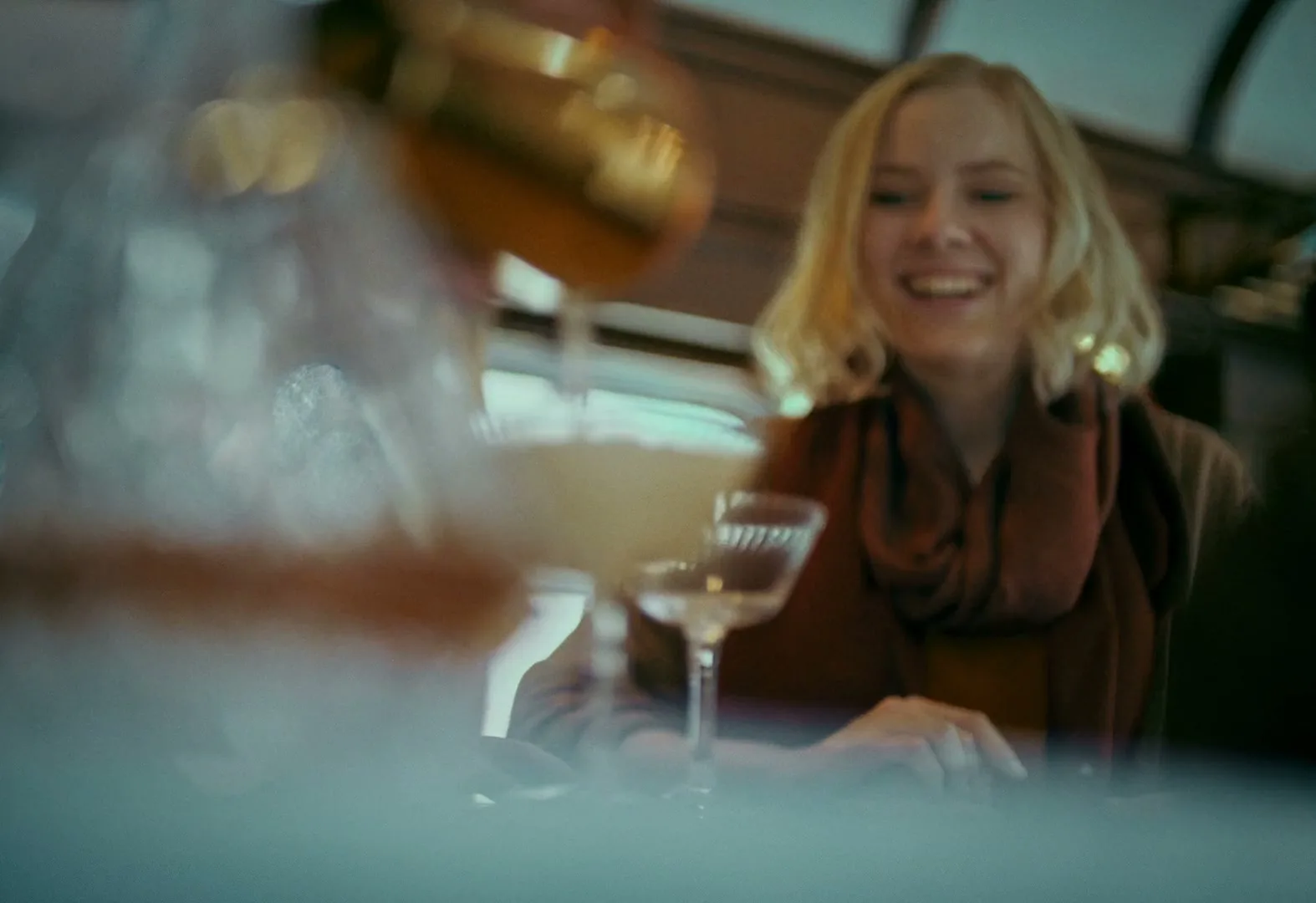 A blonde woman in a red scarf smiles happily as a bartender pours a drink into a glass in front of her on a train.