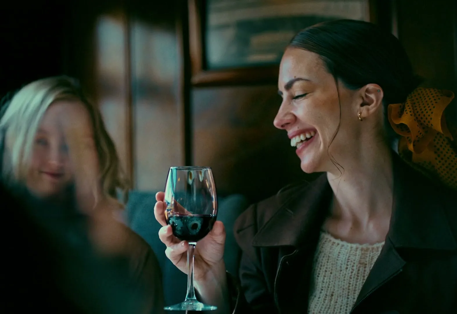 A woman with dark hair and a yellow scarf smiles broadly, holding a glass of red wine, while another woman with blonde hair is blurred in the background.