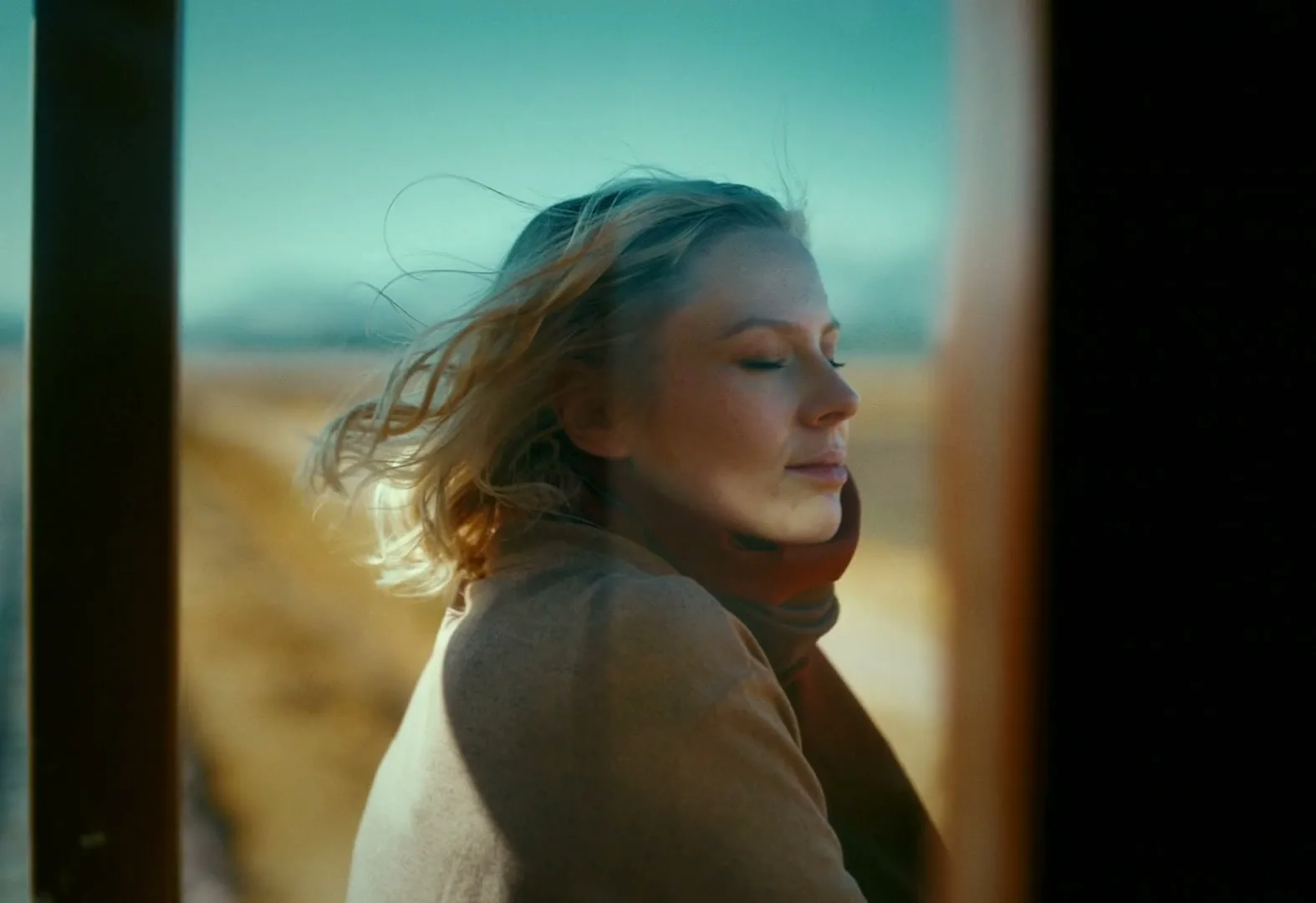 A young woman with blonde hair and a brown scarf closes her eyes, enjoying the wind on a train.